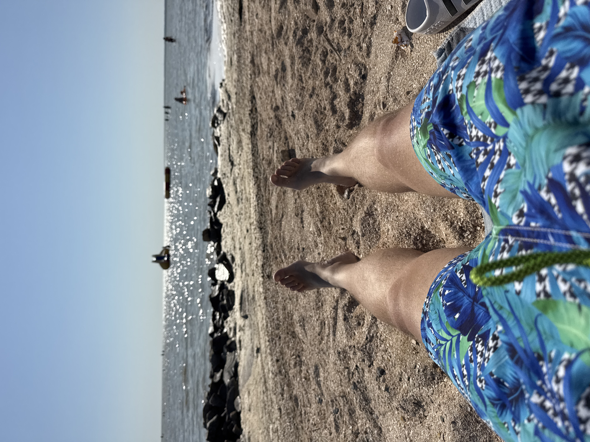 The image shows a first-person perspective of a person lying on a sandy beach, facing the sea. The person is wearing blue and green patterned swim trunks. Their bare legs and feet are visible in the foreground. The beach is covered in sand and small shells. In the background, the sea is visible with the sun reflecting off the water, creating a shimmering effect. There are several indistinct figures in the water, some appearing to be on inflatable objects. A rocky shoreline is visible separating the beach from the sea. The sky is clear and blue. A white sandal with black stripes is visible on the right side of the image. The overall scene depicts a leisure activity at a beach, likely during daytime. Given the context, this scene is likely located in Yeysk, Russia, a coastal city known for its beaches along the Sea of Azov.