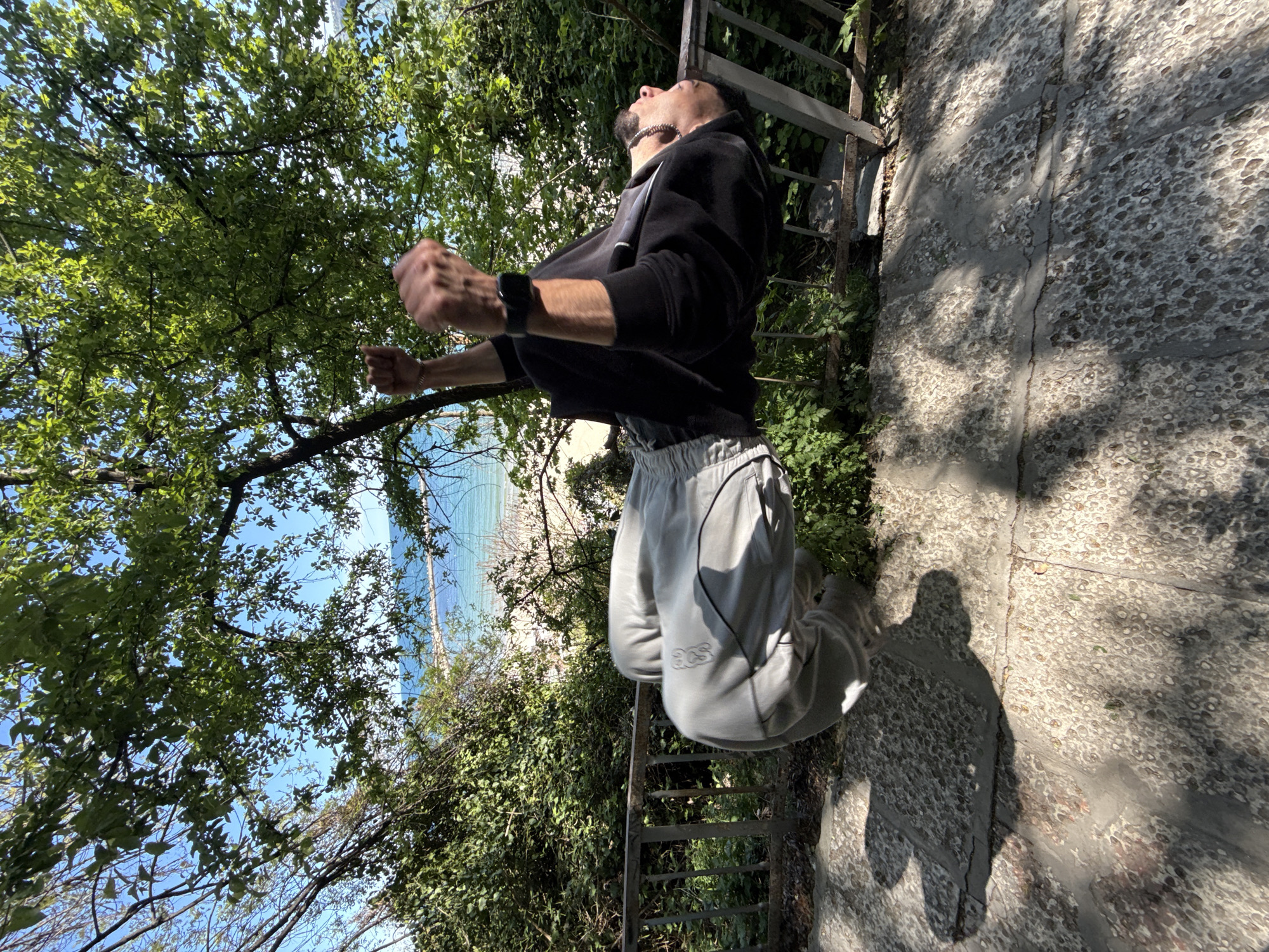 The original image is notably rotated counter-clockwise, capturing a man in mid-air during a dynamic jump outdoors. He is shown in profile, looking upwards with his mouth slightly open and both fists clenched, conveying a sense of exhilaration or effort. The man wears a black hoodie, light grey sweatpants with the text "acs" visible on his left thigh, a dark-colored watch on his left wrist, and a silver-toned chain around his neck.
The setting is an outdoor, natural environment, characterized by abundant green foliage from trees and bushes that frame the scene. Through the gaps in the greenery, a clear blue sky is visible, and in the distance, a glimpse of the sea and a sandy beach can be seen, indicating a coastal location. A concrete path or steps are visible in the foreground, along with a metal railing. Bright, direct sunlight casts long, distinct shadows on the ground, suggesting it is daytime with clear weather. The image captures a moment of energetic activity in a scenic, coastal landscape, likely in Varna, Bulgaria.