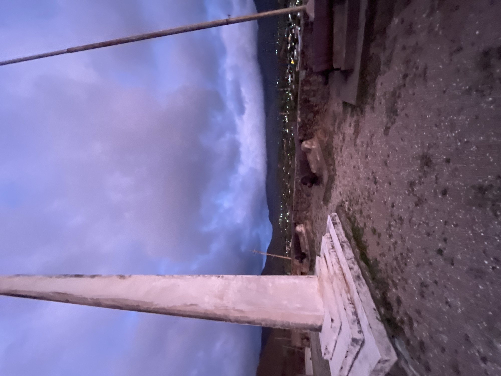 The image captures an outdoor scene at dusk, featuring a monument and cannons against a dramatic sky. In the foreground, a tall, light-colored obelisk stands to the left, mounted on a multi-tiered base. To the right of the obelisk, two cannons are positioned on raised platforms. The ground is covered with gravel and scattered debris. In the middle ground, a low stone wall runs horizontally, with more cannons visible on its surface. Beyond the wall, a town or village is nestled in a valley, with buildings illuminated by lights. In the background, dark mountains rise against a sky filled with heavy, purple and blue clouds, suggesting approaching weather or the end of daylight. The scene appears to be a historical site or a viewpoint overlooking the city of Santa Ana, Venezuela, likely Fort Cumaná due to the presence of historical cannons and the elevated position overlooking the town. No people are visible in the image.