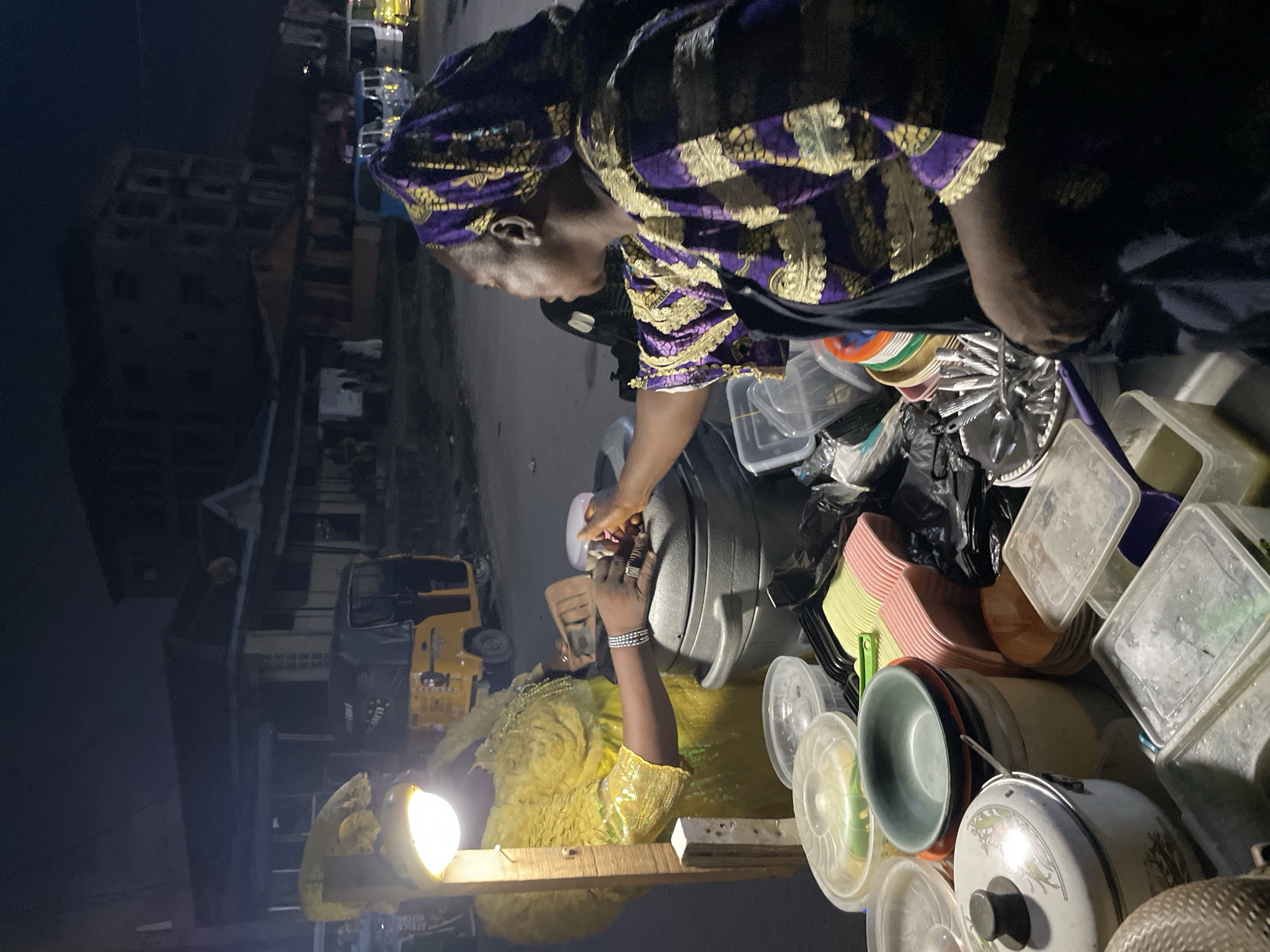 The scene depicts a woman in traditional purple and gold attire attending to a customer near a food stall at night in Ado, Nigeria. The customer, wearing a gold dress, is receiving food from a large, gray metal pot. Various plastic containers, dishes, and utensils surround the stall. A bright light illuminates the stall, and in the background, buildings and vehicles, including a yellow tricycle taxi, are visible.