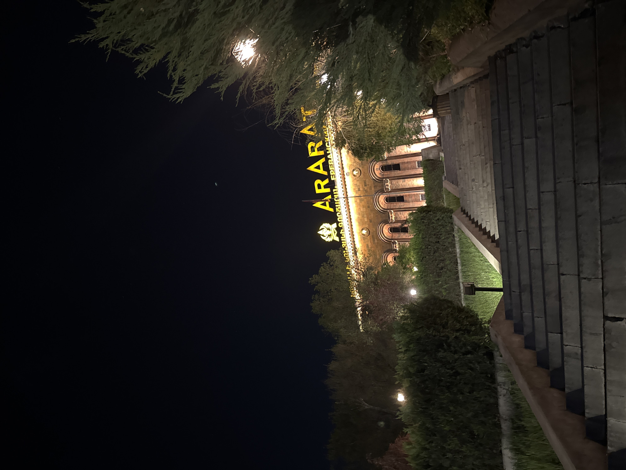 At night, a building is illuminated by lights, set against a dark sky. The building features the word "ARARAT" in large, yellow, vertically-oriented letters above its entrance, accompanied by an emblem. The architecture includes arched windows and a flat roof. Vegetation and trees obscure parts of the building. To the right of the building, a stone staircase ascends out of view. This building is the Ararat Brandy Factory in Yerevan, Armenia.