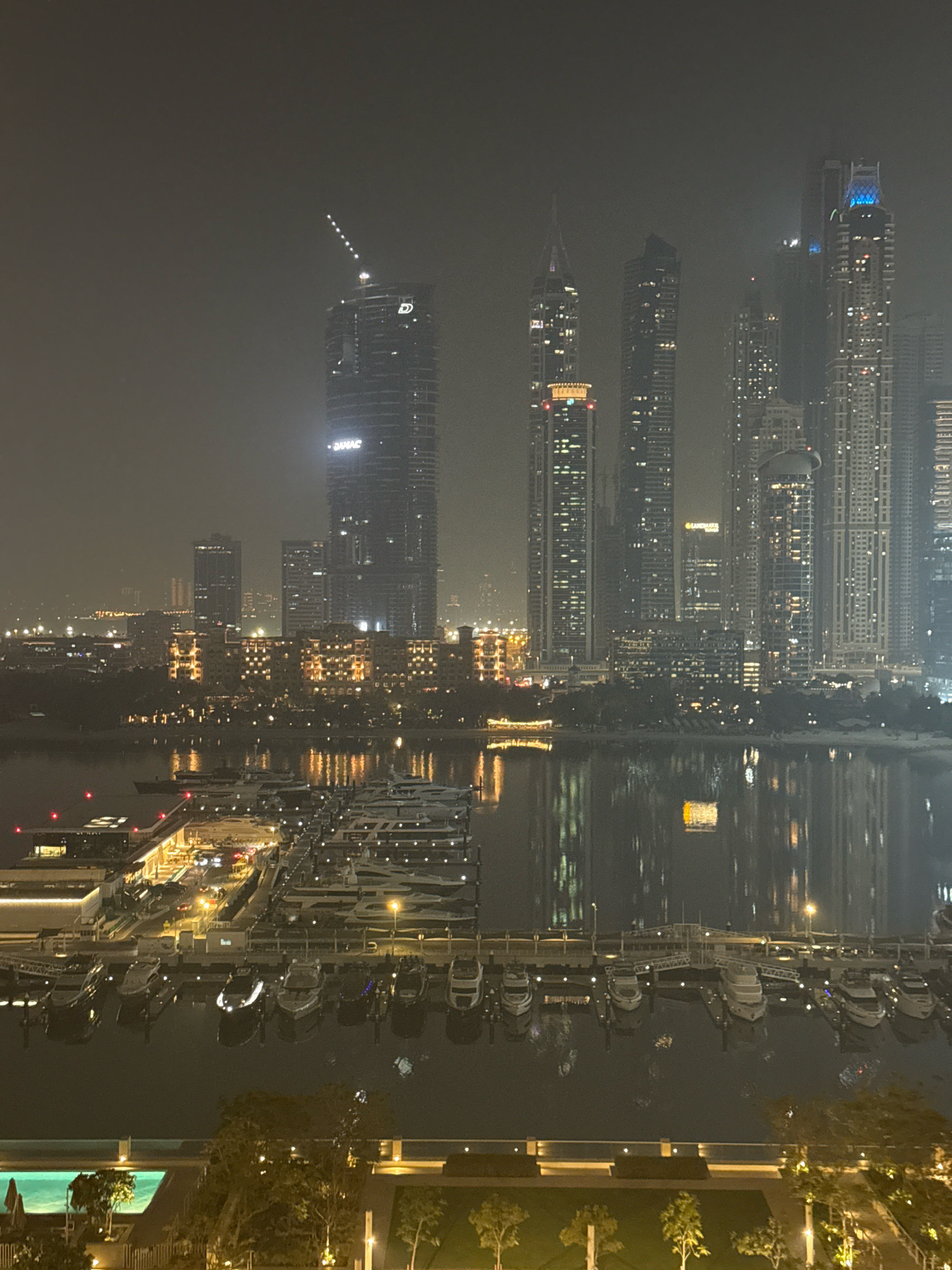 This image captures a nighttime view of the Dubai Marina in Dubai, United Arab Emirates. The scene is dominated by a collection of modern skyscrapers, illuminated by numerous lights, suggesting an urban environment. In the foreground, a marina is visible, filled with numerous yachts docked side-by-side. The water of the marina reflects the lights of the city, creating a shimmering effect. The overall atmosphere appears to be slightly hazy, possibly due to nighttime conditions or atmospheric conditions. A swimming pool and surrounding greenery are also visible in the lower portion of the image, indicating a residential or resort area. No people are clearly discernible in the image.