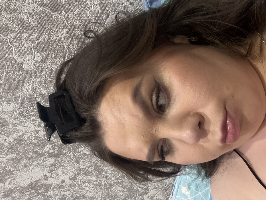 A woman lies on her back on patterned bedding. She has dark hair and fair skin. A black hair clip is fastened in her hair near her forehead. She wears makeup, including eyeshadow. Her gaze is downward. A thin black strap is visible near her shoulder.
