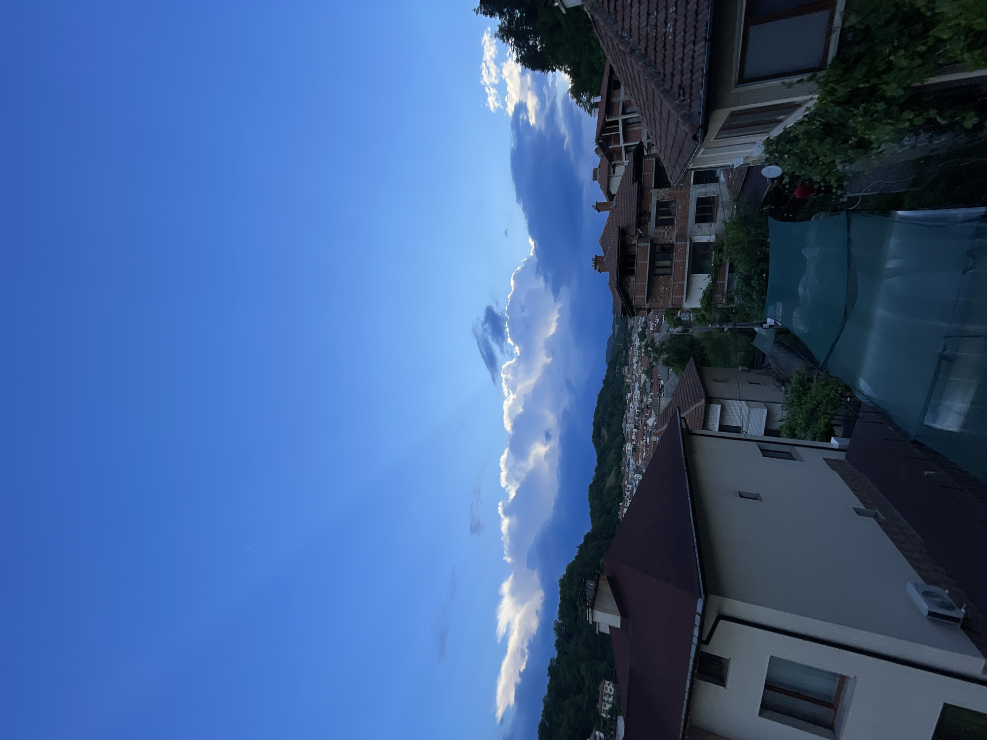 This is a vertical, low-angle shot of a sky and a hillside town. The sky is a deep blue, with wispy white clouds forming a dramatic line across the frame. Sunlight streams from the left, illuminating the edges of the clouds and casting rays across the sky. Below the clouds, the town of Zlatograd, Bulgaria, is visible, nestled on a green, tree-covered hillside. The buildings are a mix of traditional tile-roofed houses and more modern structures. The scene appears to be set in the late afternoon or early evening, given the angle of the sun and the soft lighting. There is no text visible in the image. The overall mood is peaceful and serene.