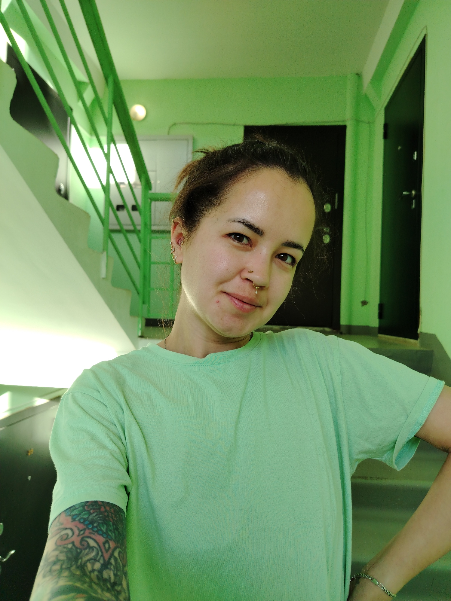 A woman stands in the stairwell of an apartment building. The stairwell is painted light green. The woman is wearing a light green t-shirt and has a tattoo on her left arm. She has brown hair pulled back in a bun, and her left ear has multiple earrings. The woman has piercings in her nose. In the background, there are black doors, stairs, and a green metal railing. The city and country are not identifiable in this image.
