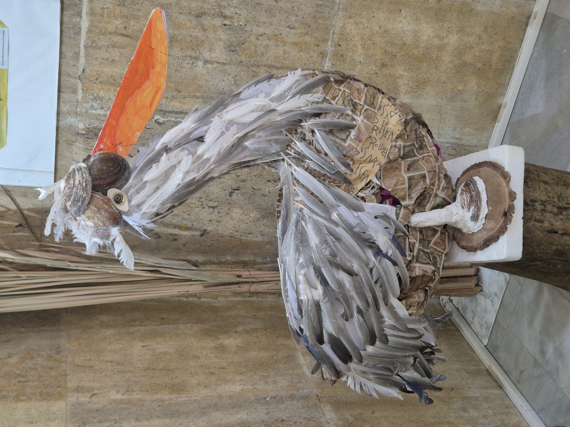 The image shows a hanging sculpture of a bird, likely a waterfowl, located at the Srebarna Lake visitor center in Srebarna, Bulgaria. The sculpture is made of various materials including feathers, shells, an orange paper cutout resembling a beak, and pieces of fabric. The bird's head is constructed from seashells and has an orange paper beak. The neck and body of the bird are draped with feathers, some gray and some white. The body's base appears to be wrapped in a patterned fabric with inscriptions. The sculpture is mounted onto a white foam board to a piece of driftwood. The background consists of a light brown textured wall. Also visible is a straw structure to the side of the bird.