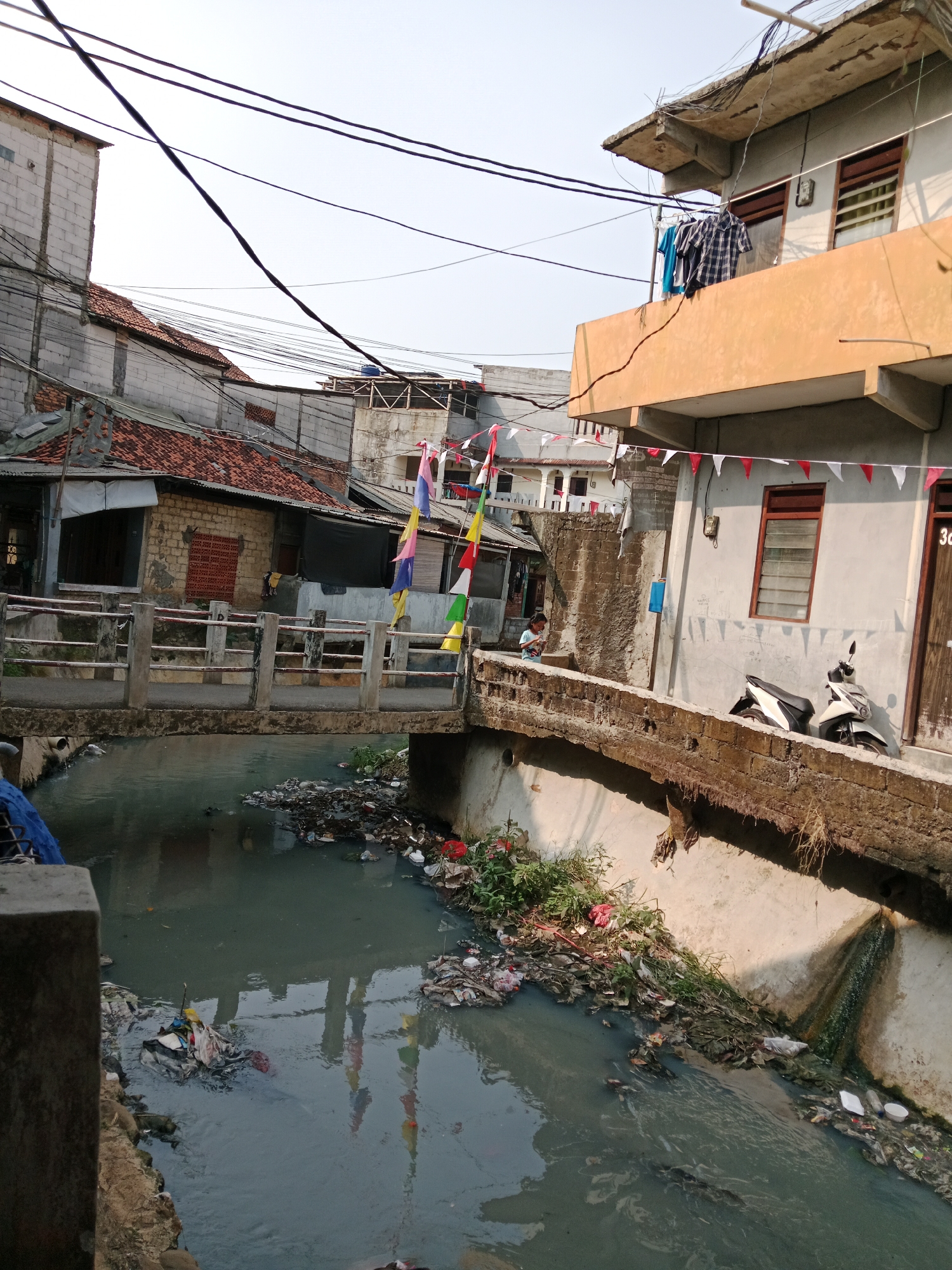 A wide shot captures a canal in an urban area of East Jakarta, Indonesia, choked with debris and refuse. The murky water reflects the surrounding buildings and sky. On the canal's banks, trash is piled high, consisting of plastic bags, wrappers, and other discarded items, with some vegetation growing amongst the litter. A concrete bridge spans the canal, its railings weathered and cracked.
On the bridge, a young girl stands near the left side, looking down at the canal, seemingly unaware of the photographer. Further to the right, on the embankment bordering the canal, two scooters are parked, one white and the other black and white. The buildings flanking the canal are multi-story structures, some appearing weathered and in disrepair. Clothes hang from lines on balconies. Decorative triangular flags, red and white, are strung between buildings, suggesting a festive occasion or national celebration. Electrical wires crisscross the sky above the scene, a common sight in urban Indonesia. The overall impression is one of neglect and environmental pollution in a densely populated urban setting.