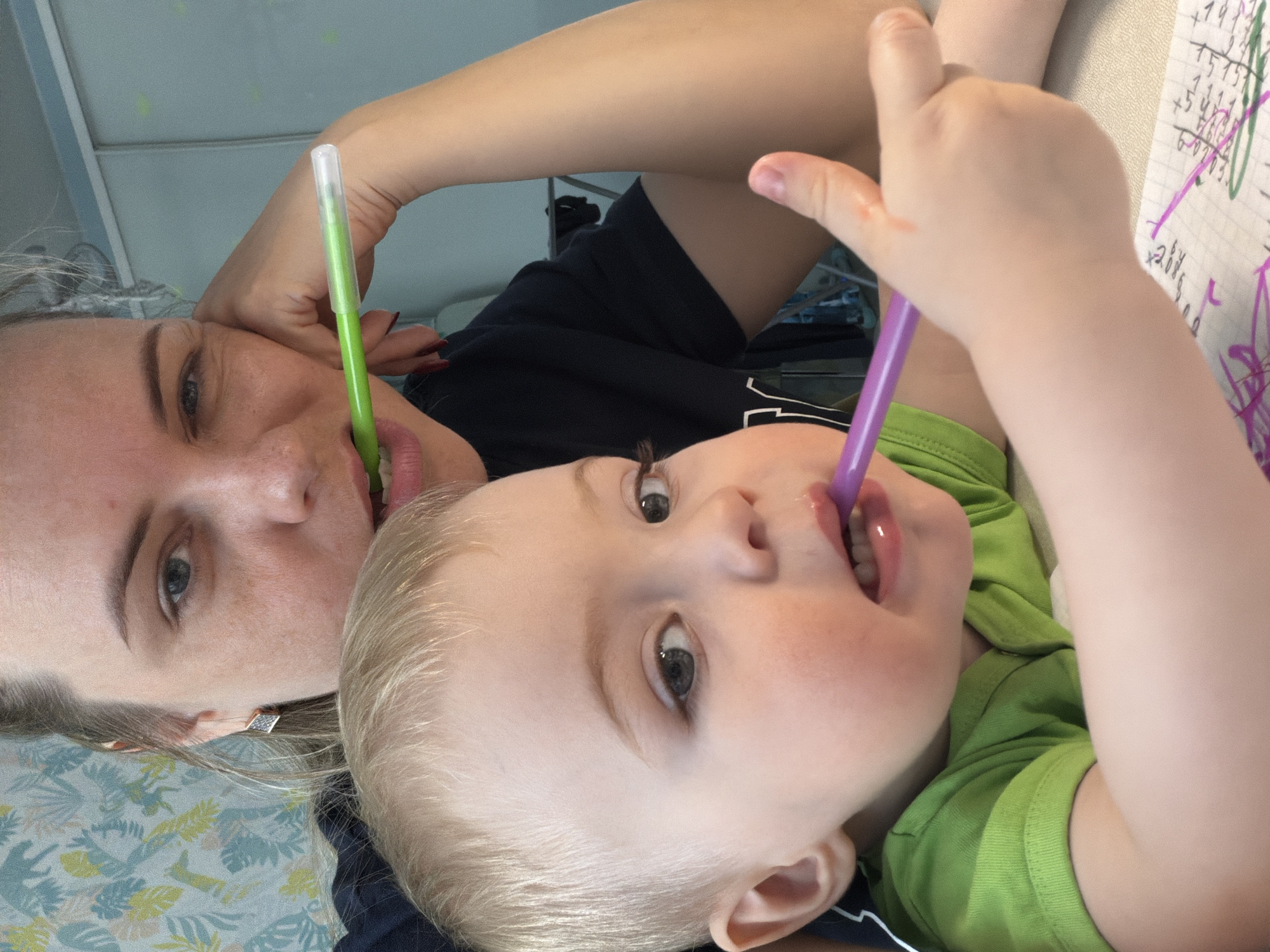 A selfie taken in Votkinsk, Russia shows a woman and a young child lying on a surface, possibly a bed or floor. Both individuals are holding pens in their mouths. The woman has light skin, dark hair pulled back, and is wearing a dark shirt. The child has fair skin, blonde hair, and is wearing a green shirt. A piece of paper with math problems is also visible in the background.