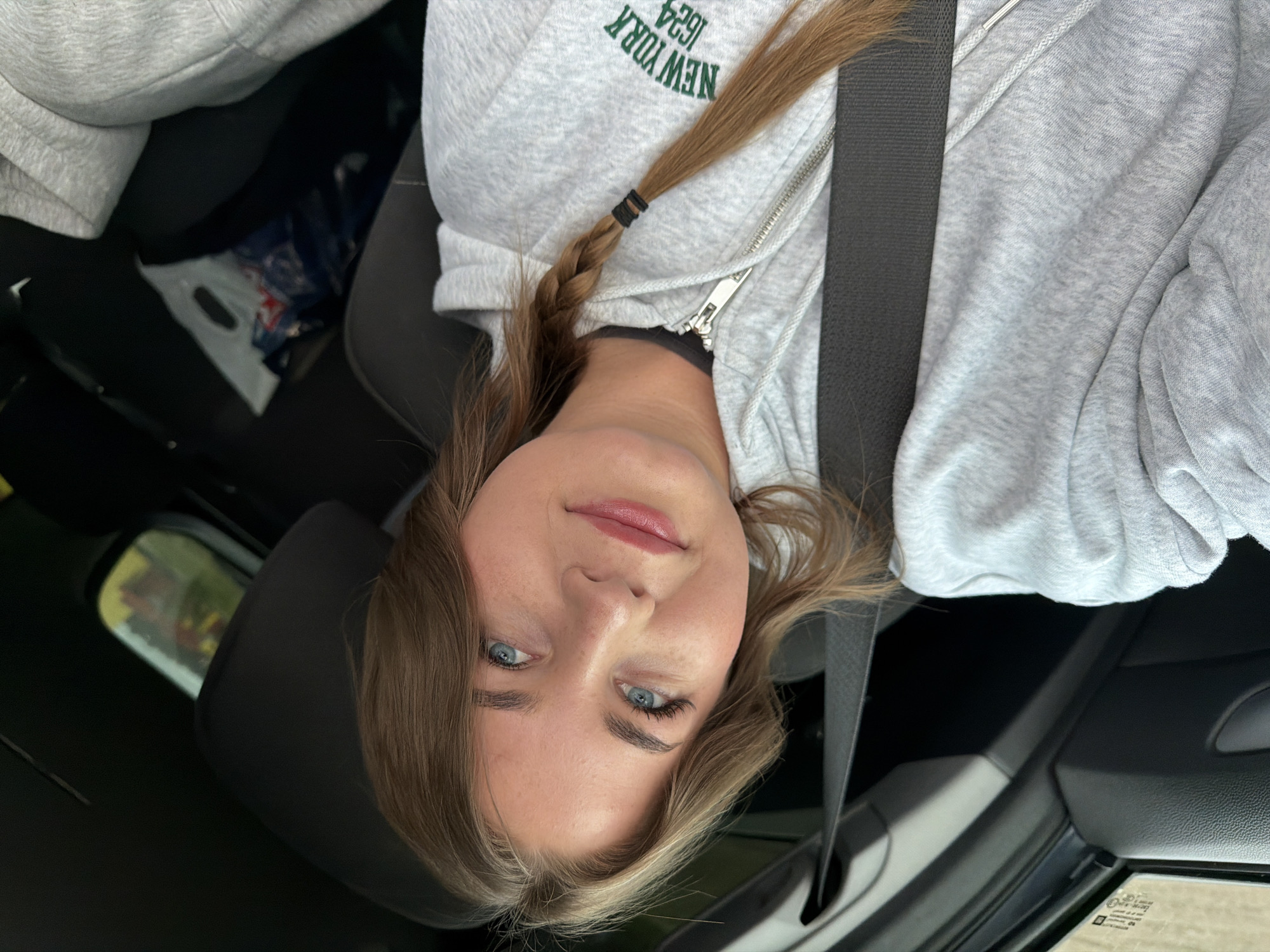 A young woman with light brown hair braided on one side and blue eyes is sitting in the driver's seat of a car, wearing a light gray zippered hoodie with "NEW YORK 1624" printed on it. She is wearing a seatbelt. The interior of the car is visible, with black seats and some items in the passenger seat. The background through the car window suggests an outdoor environment. There is no indication of the location being Chaikovsky, Russia.