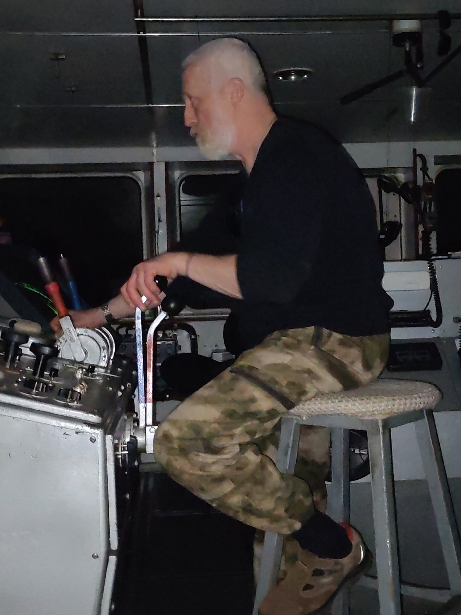 A medium shot captures a white-haired man with a beard seated on a stool, operating machinery within an enclosed space. The man is dressed in a dark long-sleeved shirt and camouflage pants. His left hand is positioned on a lever, while his right hand is grasping a black handle. He is wearing a watch on his left wrist. The machinery appears to be gray and metallic, featuring various levers, dials, and switches. In the background, there is a window showing a dark exterior and equipment, including wires and a microphone. The stool has a gray metal frame with a woven seat. The location is Unknown, Unknown.