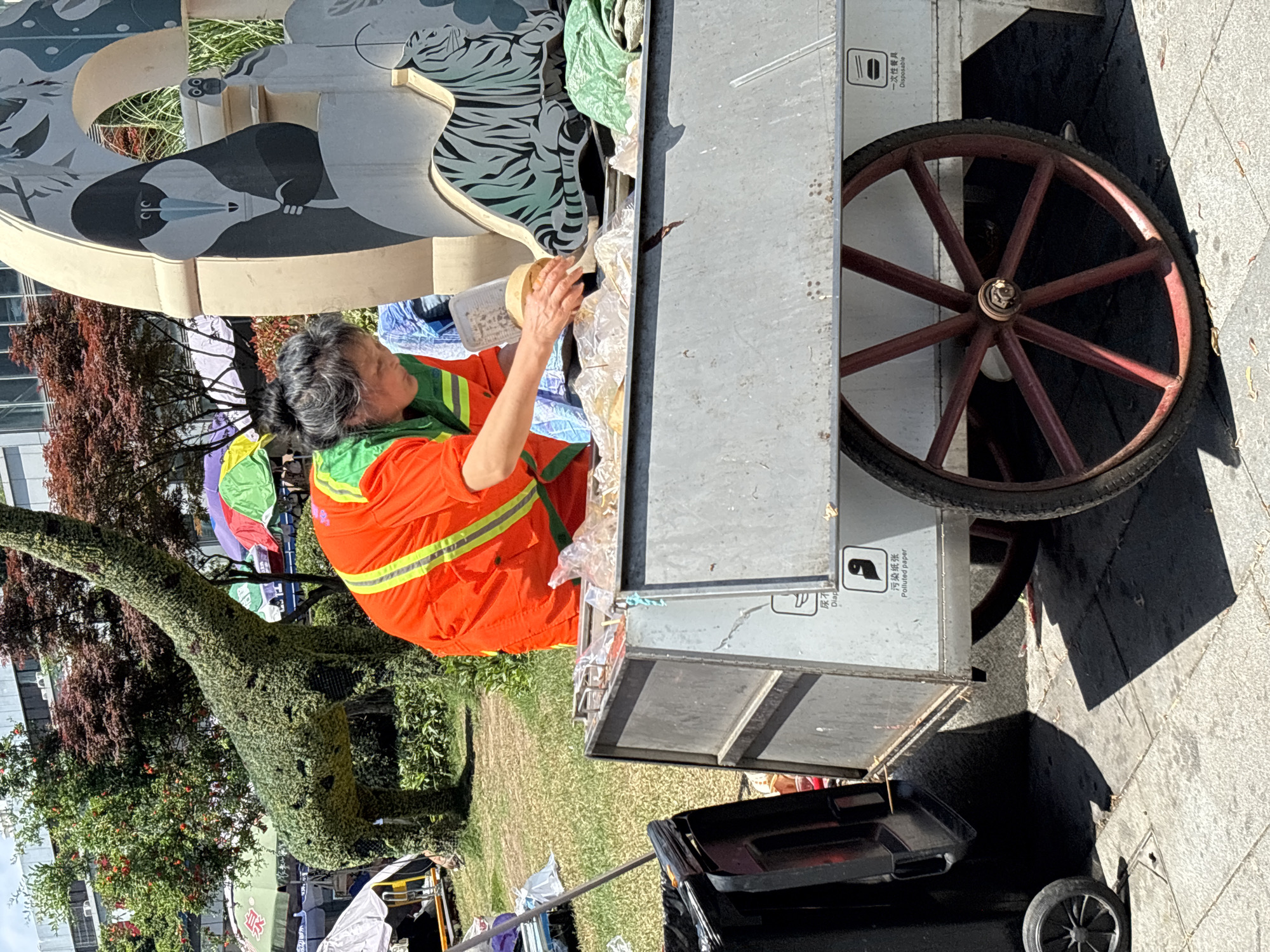 In Jiulongpo District, China, an elderly woman in an orange and green high-visibility uniform stands at a light gray cart with a large, dark red spoked wheel, appearing to sort through items on the cart's surface. The cart is parked on a paved area adjacent to a grassy lawn featuring a dinosaur-shaped topiary. The cart's side displays icons and text in Chinese, possibly related to disposable products. A black waste bin is positioned near the base of the cart. Above the woman and the cart, there is a decorative structure featuring animal illustrations, including a penguin-like bird and a zebra. The background includes trees and buildings, indicating an urban park or garden setting.