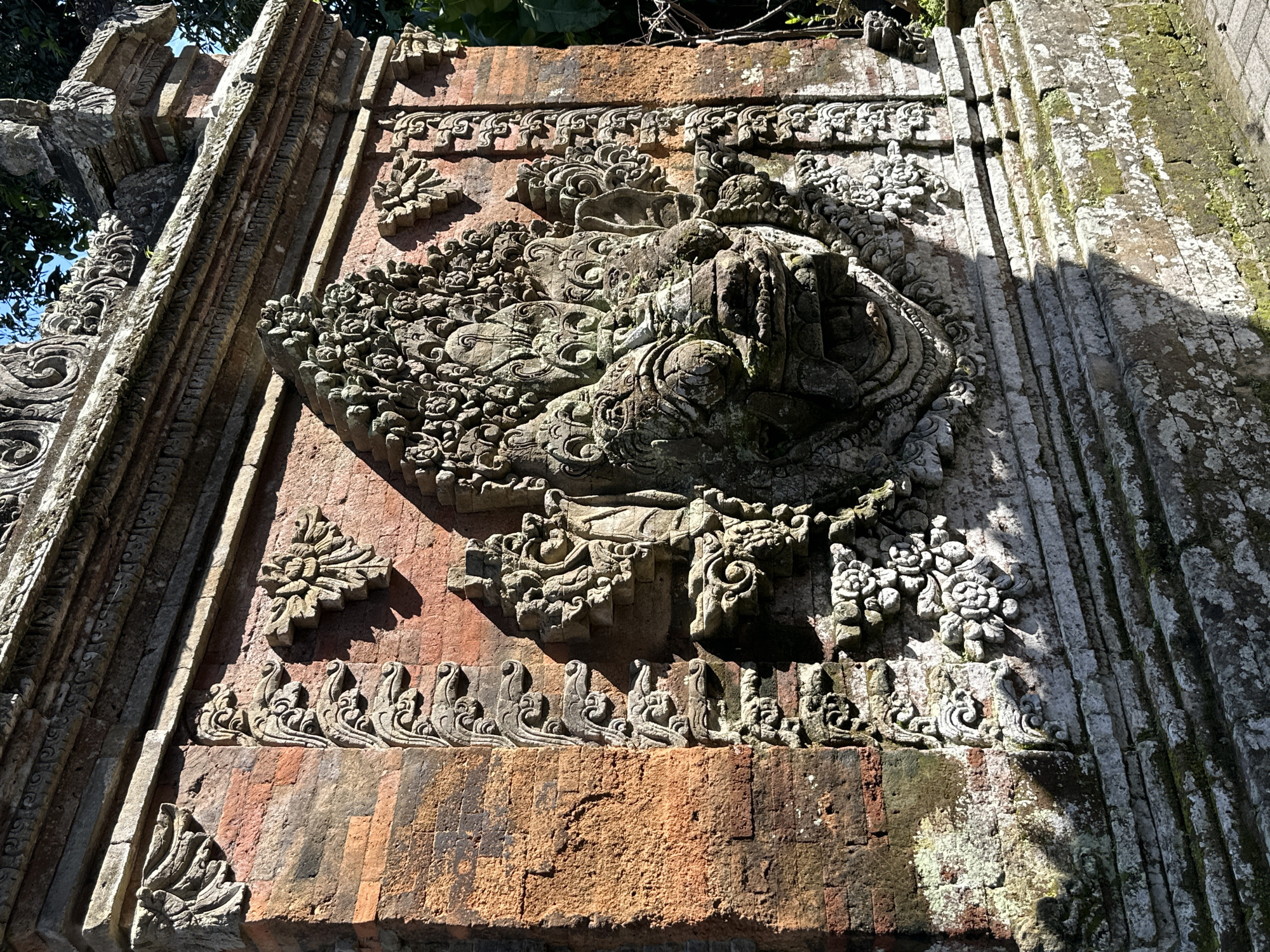 The image depicts a close-up view of an intricately carved stone face integrated into a red brick wall, likely a section of a temple or historical structure in Ubud District, Indonesia. The stone carving is detailed, featuring a fierce expression with bulging eyes, sharp teeth, and elaborate headwear adorned with floral and scroll motifs. The brickwork exhibits signs of age, with some areas showing moss and weathering, suggesting it is an outdoor structure exposed to the elements. The sunlight casts strong shadows, highlighting the three-dimensional textures of the carvings and the brickwork. The background is blurred, with hints of green foliage and a clear blue sky visible at the top. No people or contemporary elements are visible in the frame.
