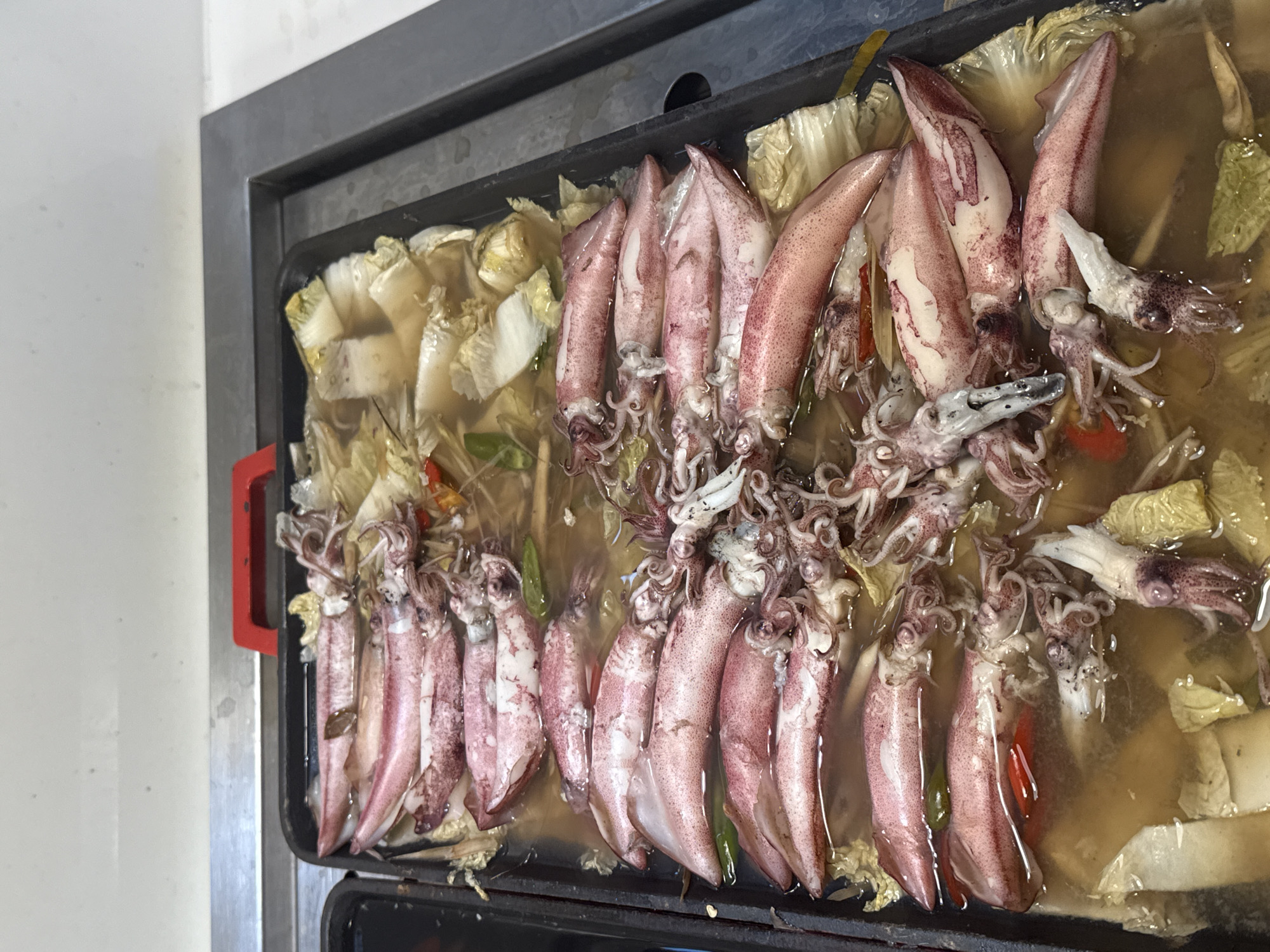 A close-up, high-angle, eye-level shot shows a black metal rectangular tray filled with cooked squid and napa cabbage in a light brown broth. The squid are arranged in two rows, with their bodies submerged in the broth and their tentacles visible above the surface. The squid are pinkish-white with red speckles and have visible eyes and tentacles. The napa cabbage leaves are pale green and white, some appearing wilted in the broth. Red chili slices and green pepper pieces are scattered throughout the tray. A red handle is visible on the far left side of the tray. The tray is placed on a gray metal surface, likely a buffet or serving station. The background is blurred and appears to be a wall or counter. The scene is consistent with a food establishment in Nha Trang Ward, Vietnam, possibly a buffet or hot pot restaurant.