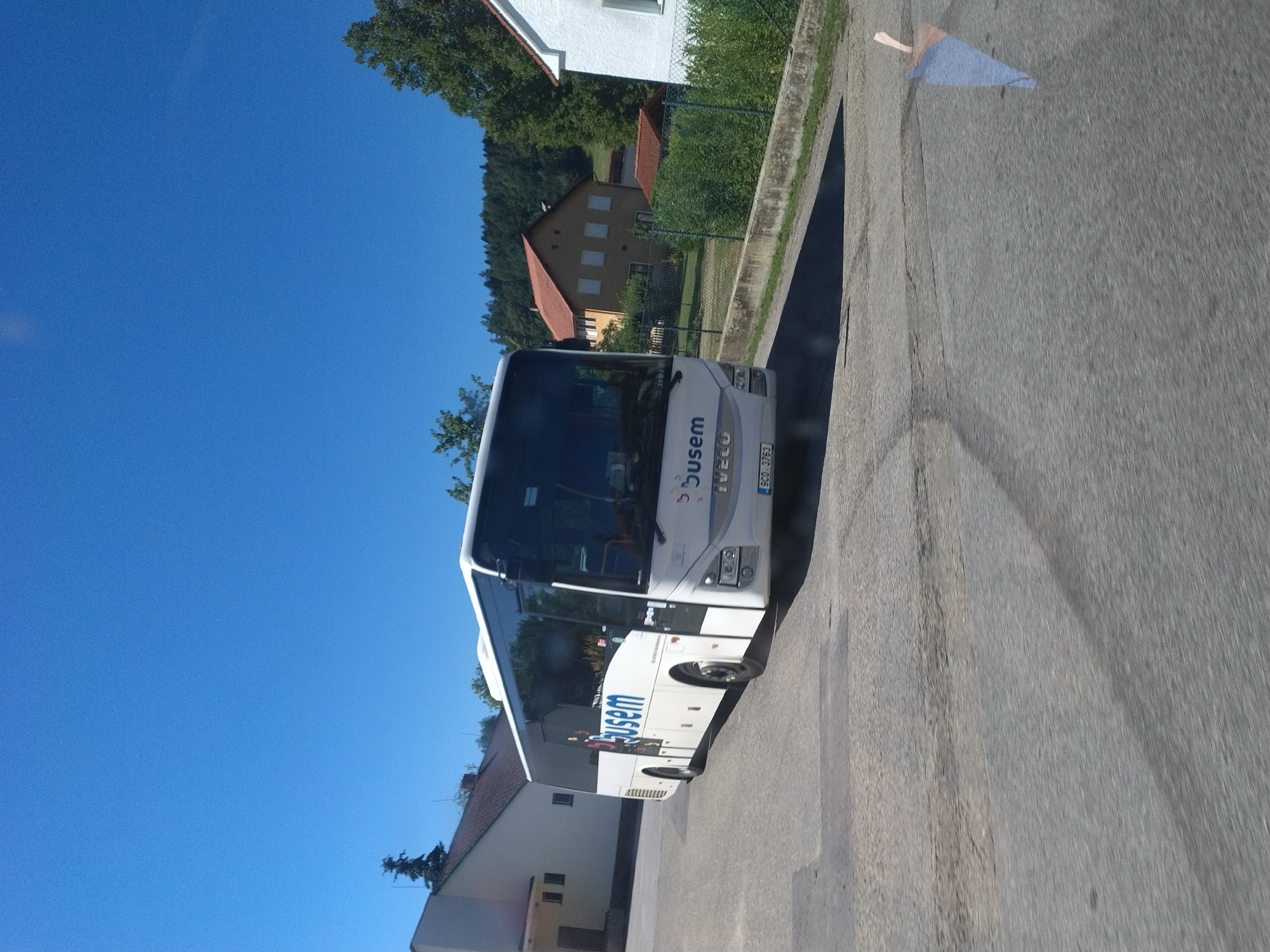 The image depicts a white "Busem" bus with "Iveco" branding on the front, parked on an asphalt surface. The bus is the primary subject and is positioned slightly to the left of the frame. In the background, there are several buildings, including a white building with a brown roof to the left, and a yellow building with a red roof further to the right. A fence and lush green vegetation are visible between the buildings and the road. The scene is set against a clear blue sky. The photo appears to have been taken from a moving vehicle, indicated by the slight blur in the foreground. The location is identified as Nová Ves, Czechia. There are no people visible in the image.