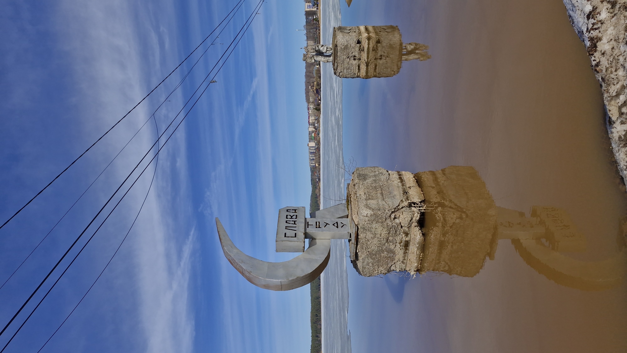 The image shows a monument in the city of Tolyatti, Russia, with the text "СЛАВА ТОЛЬЯТТИ". The monument has a large metallic crescent shape and rests on a concrete base. The image is taken from a low angle, near a body of water, which reflects the monument and the sky. Overhead power lines are visible in the upper-left corner against a blue sky with streaky clouds. The water is brownish, and a shoreline with some remaining ice is seen on the right. In the background, there is a line of trees and buildings. The photo appears to have been rotated slightly.