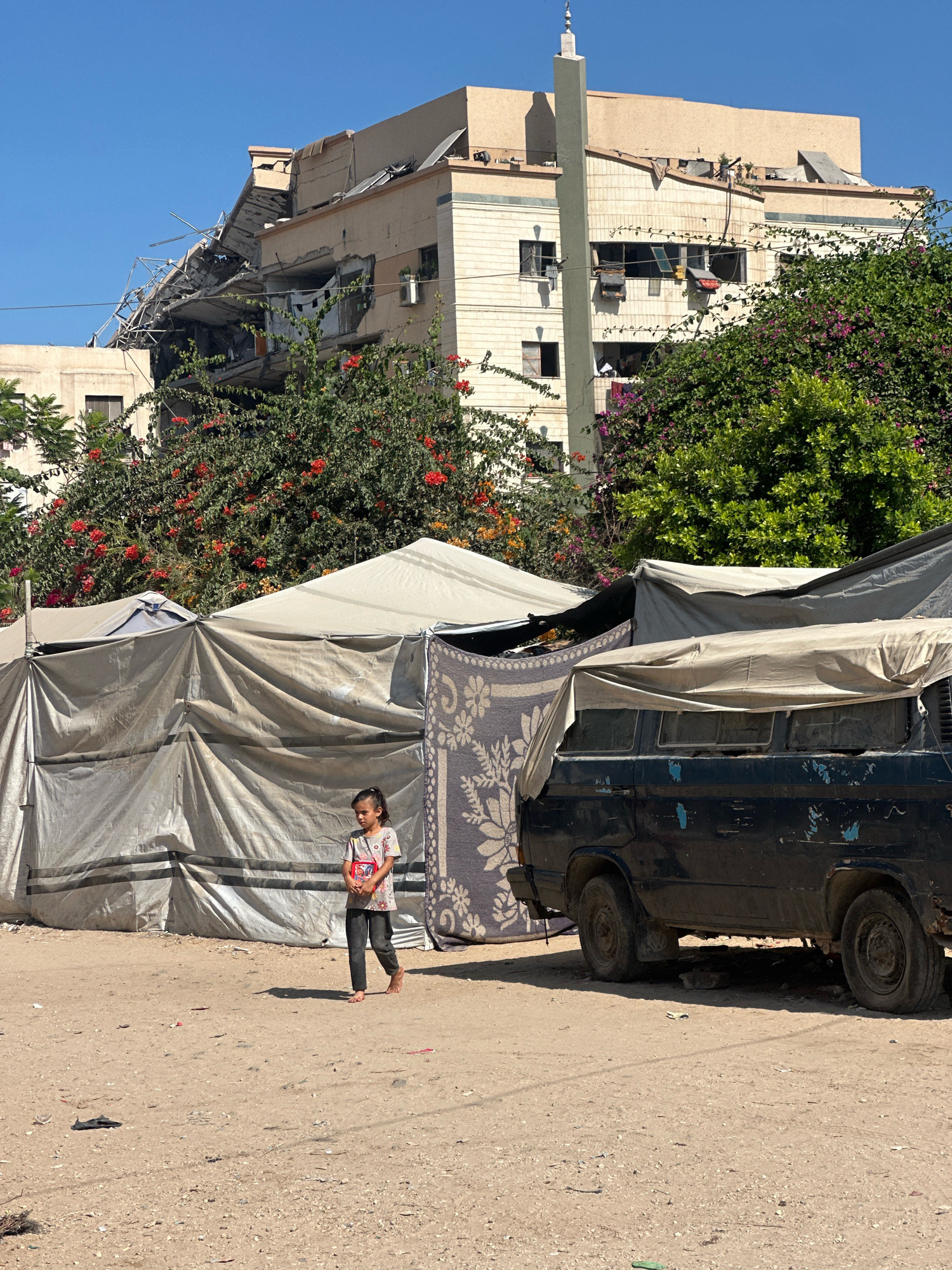 In the city of Gaza, Palestinian Territories, a young girl walks barefoot across a dirt ground. She is wearing a colorful t-shirt and dark pants, carrying a red object in her hands. Behind her, a large dark blue van is partially covered by a tarp. Further back, a series of makeshift tents are erected, constructed from large sheets of grey fabric. In the background, a building with significant structural damage is visible, with debris and exposed framework. Lush green trees and flowering plants, with red and purple blossoms, surround the damaged structure and tents. The sky is clear and blue.