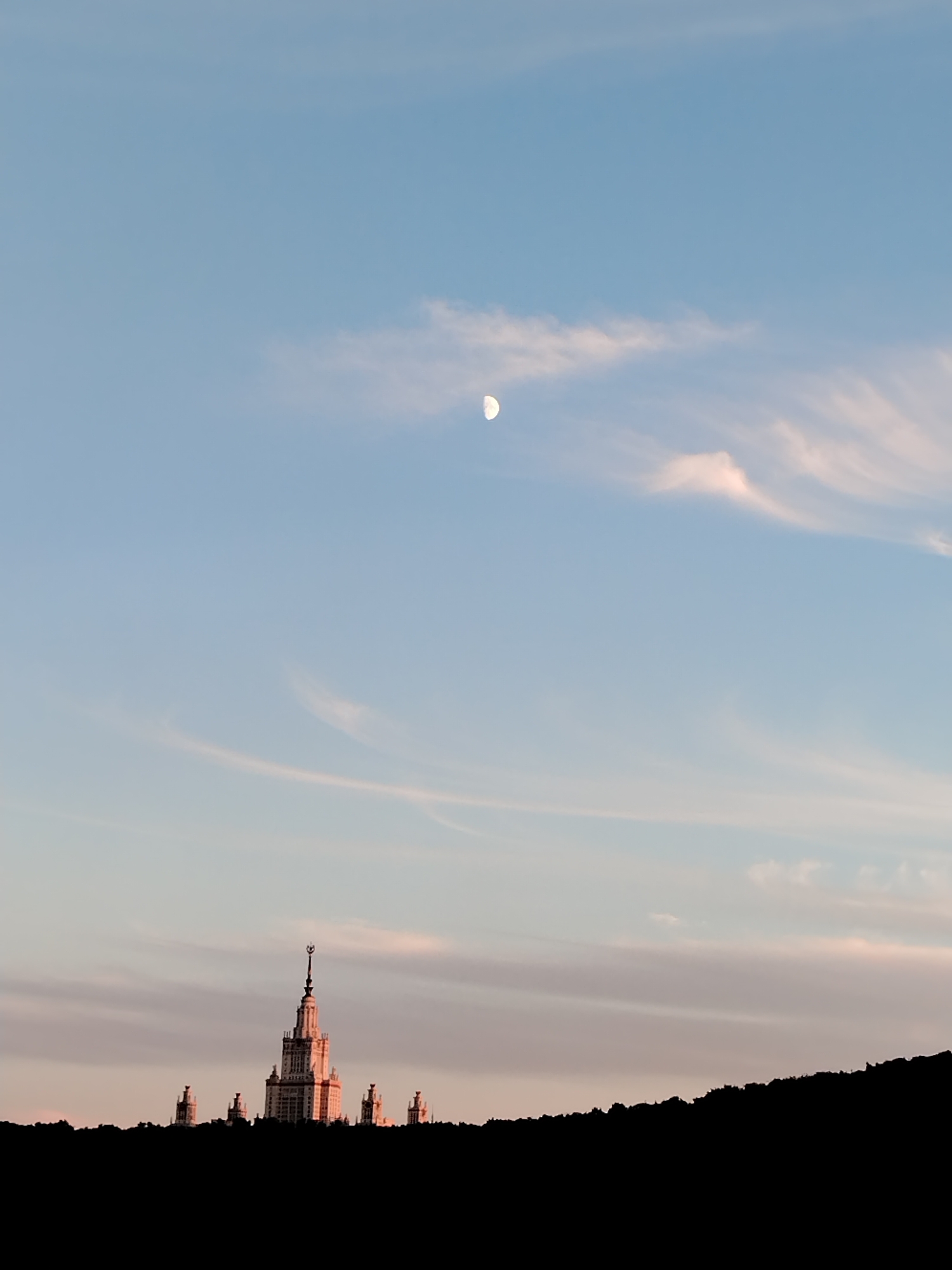 A vertical shot captures a twilight sky with a crescent moon visible high above. Wispy clouds streak across the pale blue expanse, tinged with pink and orange hues from the setting sun. In the foreground, silhouetted against the colorful sky, is the distinctive outline of a tall, multi-tiered building with spires, resembling a monumental structure like a university or government building. Several smaller, similar structures flank the main building, all appearing to be part of a complex. The entire cityscape is framed by a dark, jagged silhouette along the bottom of the image, suggesting a hill or a dark foreground element. The overall mood is serene and majestic, evoking a sense of awe at the grandeur of the architecture against the dramatic backdrop of the evening sky. The time of day is clearly dusk or twilight, indicated by the soft lighting and the presence of the moon. The weather appears clear, with only scattered, delicate clouds.