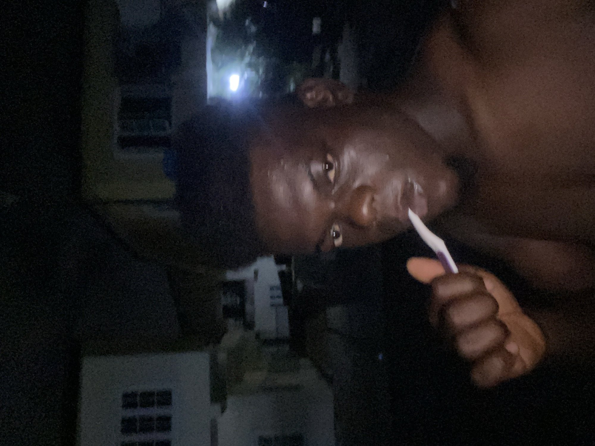 An eye-level close-up captures a dark-skinned individual with short, dark hair brushing their teeth with a white and purple toothbrush. The person's bare shoulder is visible on the right. Behind them, a partially visible building stands in the background, discernible by its windows. The location and city are unknown. The overall lighting is poor, making details difficult to discern.