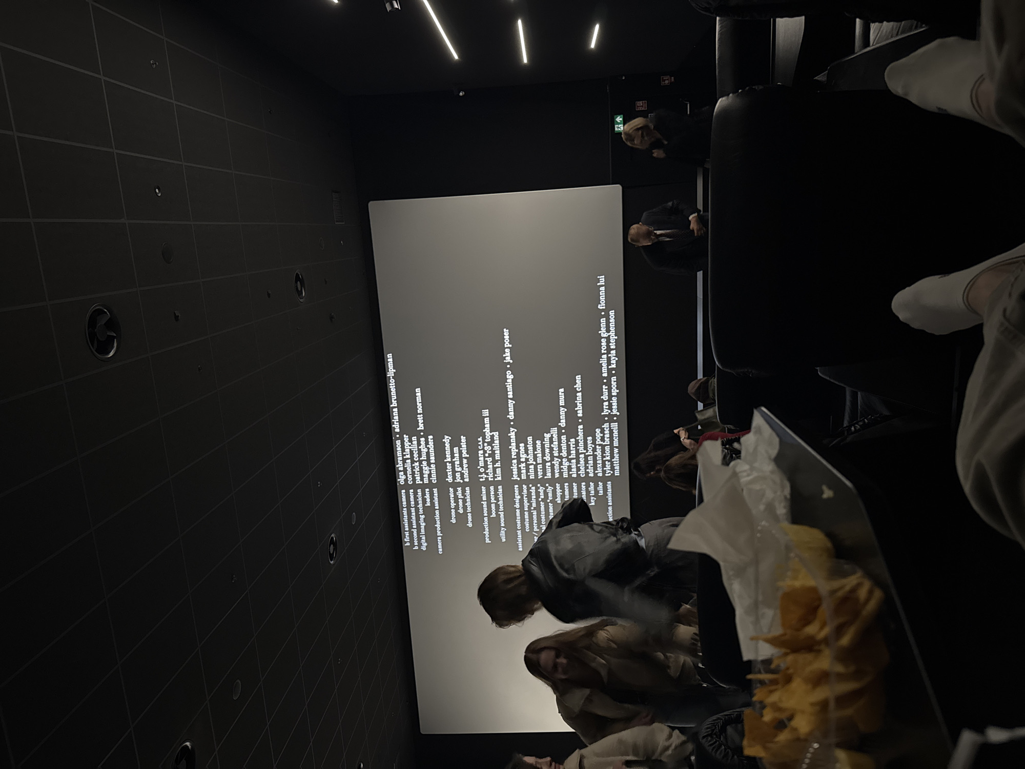 The image shows a dimly lit movie theater with a large screen displaying the end credits of a film. The environment is dark, typical of a cinema during a showing, with rows of tiered seating. In the foreground, there are several people sitting, with a container of chips or snacks prominently featured. A few others are further back, closer to the screen. The seating appears to be black leather or a similar material. The screen displays white text against a light background, and the ceiling has a few linear lights providing minimal illumination. The overall impression is of the end of a movie experience with people relaxing and potentially snacking as the credits roll.