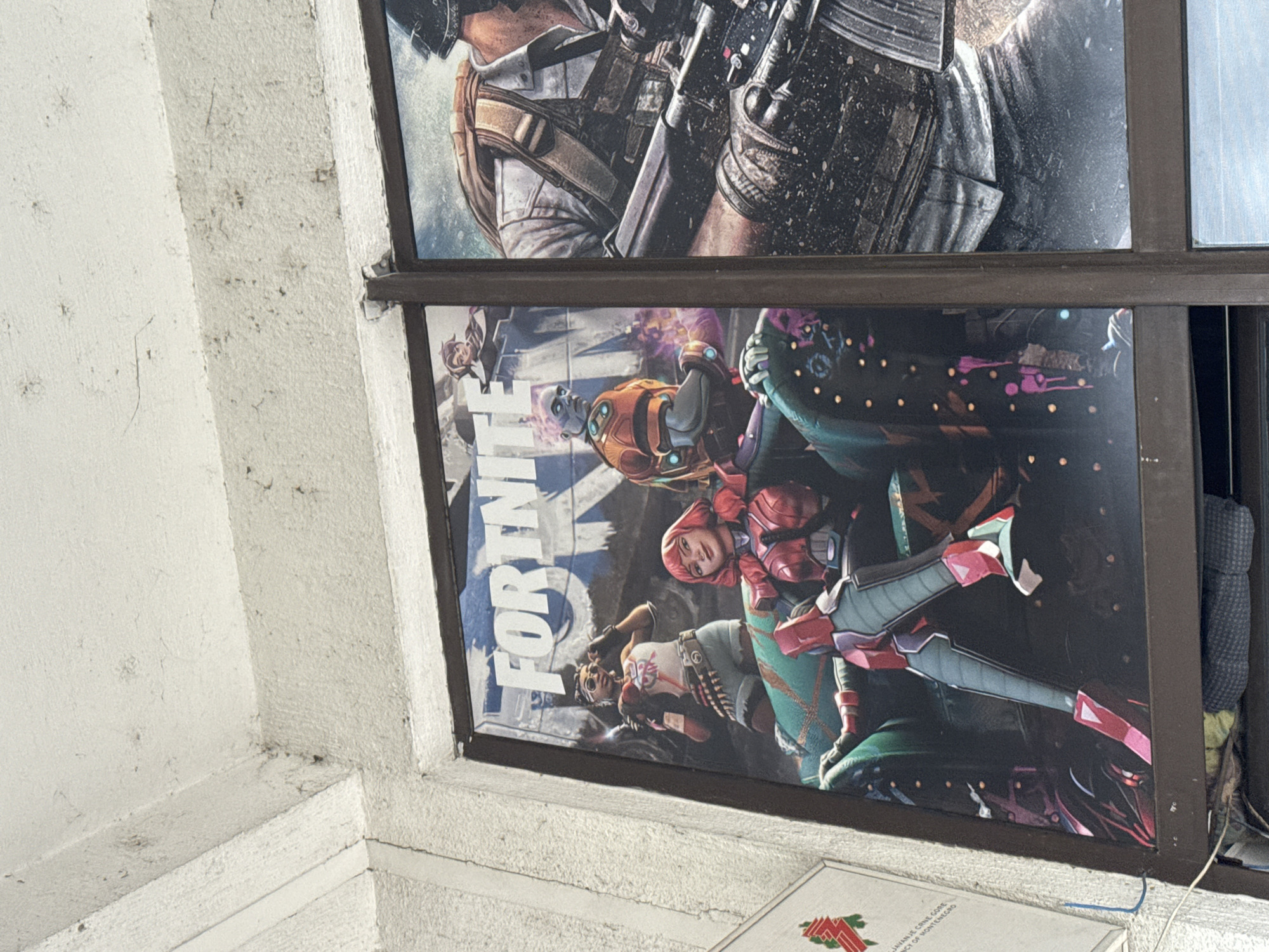The image shows a section of a window display at an establishment, likely a business or residence, in Šušanj, Montenegro. The display consists of two posters within a dark-colored frame. The upper poster features a person holding a weapon. The lower poster is a Fortnite-themed image with several characters and the game's logo prominently displayed. A portion of a wall, textured and painted white, is visible to the left of the window frame. The window is slightly ajar, revealing some items inside. A white document with red and green graphics is partially visible below the window. Wires are also present around the window.