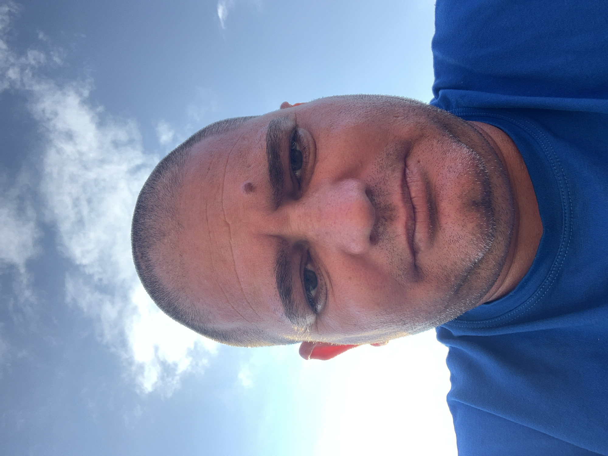 A close-up, low-angle self-portrait of a middle-aged Caucasian man with a shaved head, light stubble, and a mole on his forehead. He is wearing a blue t-shirt. The background shows a bright, blue sky with scattered clouds. The location cannot be determined from this image and there is no indication in the image that it was taken in Zetiovo, Bulgaria.