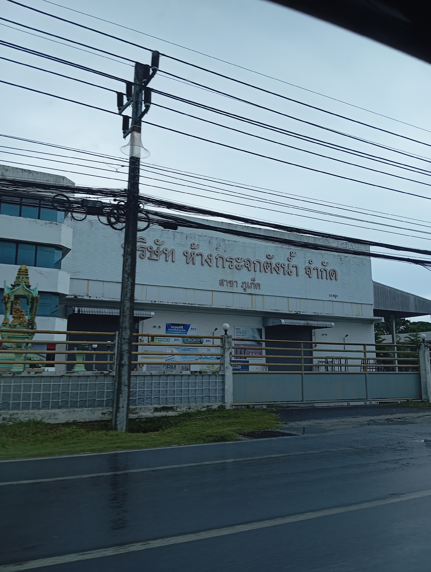 The image shows a building with a sign that reads "บริษัท ห้างกระจกน้ำ จำกัด สาขา ภูเก็ต" in Thai, indicating a company name and a branch in Phuket. The building is white with large windows and a metal roof. There is a fence in front of the building, and a road with wet pavement in the foreground, suggesting it may have recently rained. Overhead, numerous power lines crisscross the sky, supported by a utility pole. A small decorative statue is visible to the left of the building. The sky is overcast.