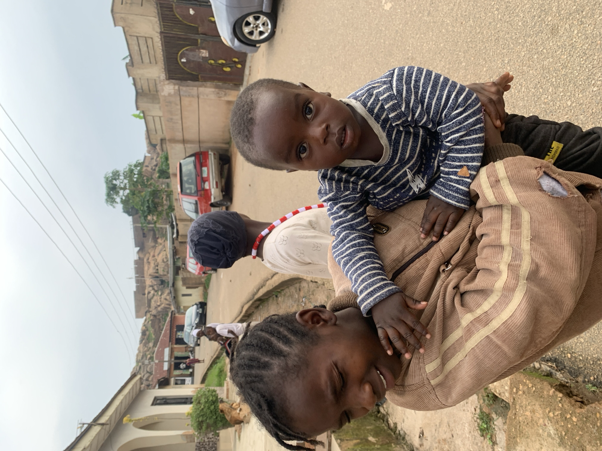 This is a close-up, vertically oriented photograph of a group of people in what appears to be a street scene in Jos, Nigeria. The primary focus is on two children, a toddler being held by an older child or adult. The toddler, dressed in a blue and white striped shirt, is looking directly at the camera with a curious expression. The older individual is wearing a brown corduroy jacket and is supporting the toddler. The faces of both individuals are visible, with the toddler's appearing brighter and more alert, while the older person's face is partially obscured and seems to be smiling or laughing.
In the background, a red vehicle and a grey car are parked on a paved surface, suggesting a residential or commercial street. Buildings with distinct architectural styles and some vegetation are also visible, hinting at the urban environment of Jos. The sky appears overcast, indicating a cloudy day. The overall mood of the image is intimate and captures a moment of interaction between the children. The clothing styles and the overall environment suggest a contemporary setting in Nigeria. No visible text is present in the image.