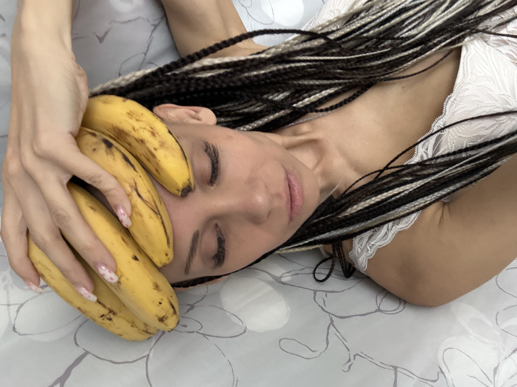 A woman lies on a bed with a light gray, patterned sheet. She has brown and white braids and is wearing a white lace top. Her eyes are closed, and she is holding three bananas on her forehead with her right hand. She has long, decorated fingernails. The location is not discernible from the image.