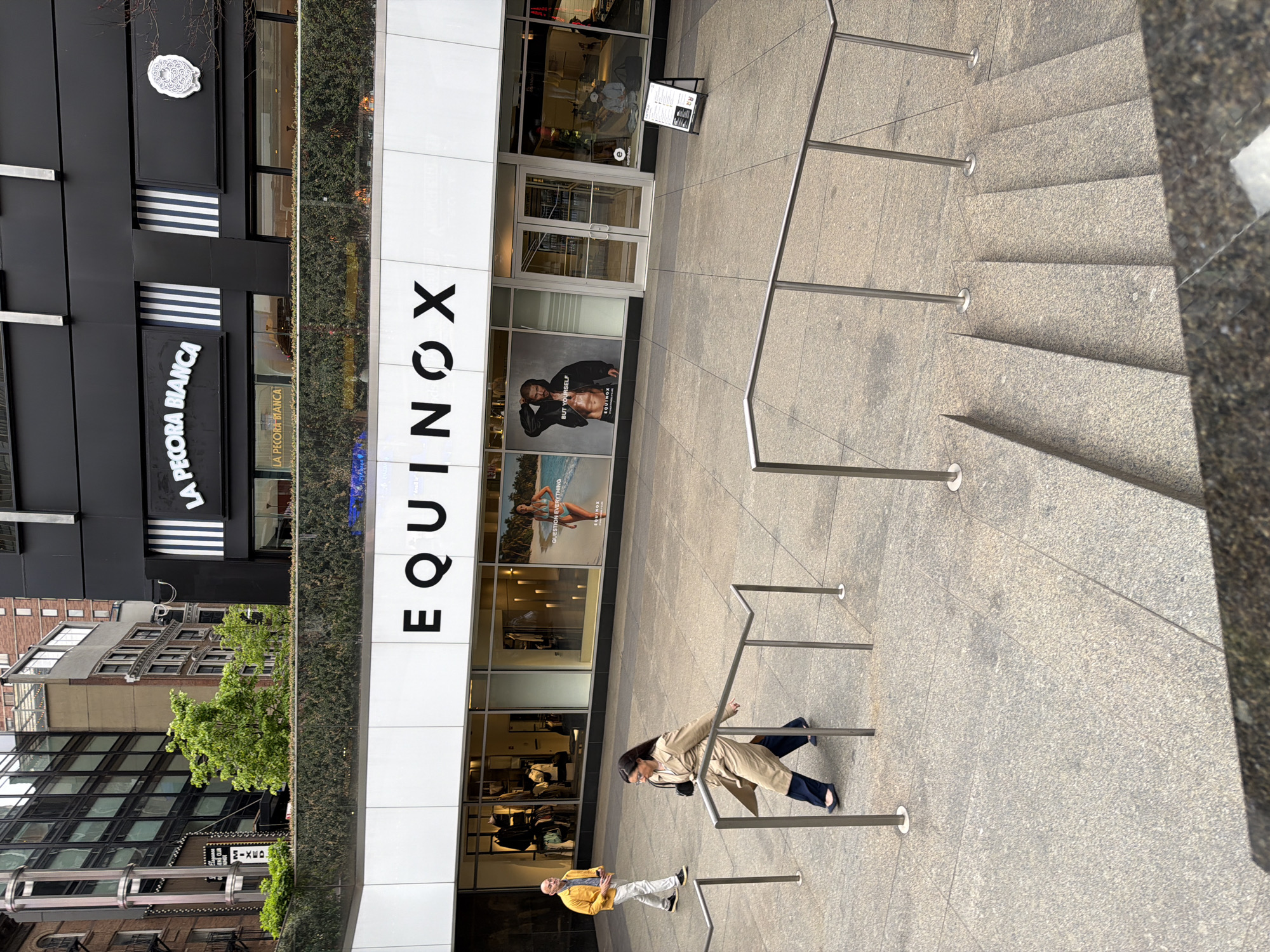 The image shows the exterior of an Equinox fitness center. To the left of Equinox, there is a restaurant called La Pecora Bianca. Two people are walking past the front of the business. A woman is wearing a tan trench coat and navy pants. The other person is wearing a yellow jacket and white pants. The building is located in New York City, New York, United States of America. There are metal handrails near a set of stairs.