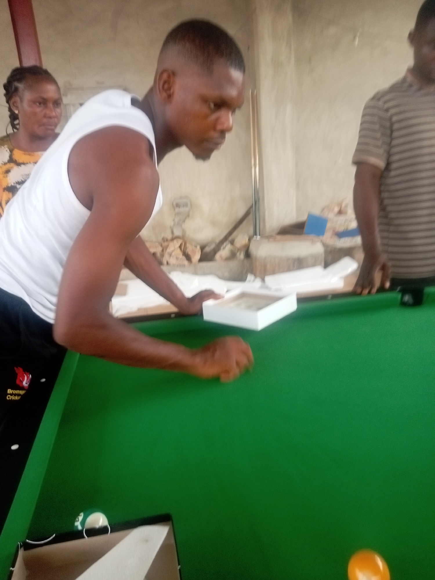 In a dimly lit indoor setting in Umuokpu, Nigeria, a man wearing a white tank top and black pants stands next to a green pool table. He is looking down and appears to be holding a small, white, square box. Two pool balls, one orange and one green and white, are visible on the table, along with a cardboard box. Behind him and to the left is a woman with braided hair, wearing a patterned dress. To the right of the man, partially visible, is another man wearing a striped shirt. The background includes rough, unfinished walls and miscellaneous objects.