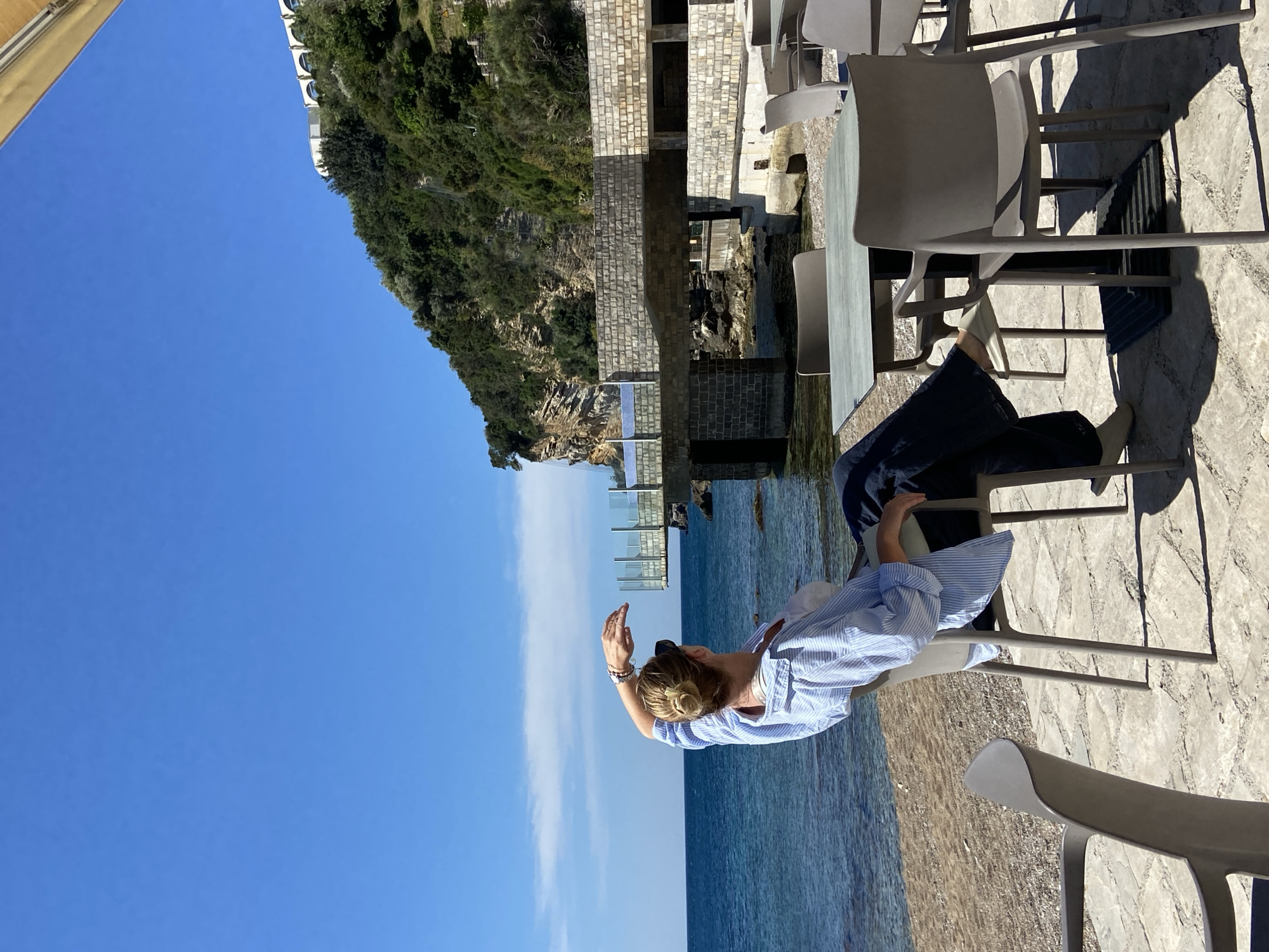 A woman in a blue and white striped shirt and dark pants is reclining in a gray chair at an outdoor cafe. The cafe is situated on a stone patio overlooking the Adriatic Sea in Budva, Montenegro. In the background, a cliff covered in lush green vegetation is visible, topped by buildings. Other gray chairs and tables are scattered on the patio. The sky is clear and blue.