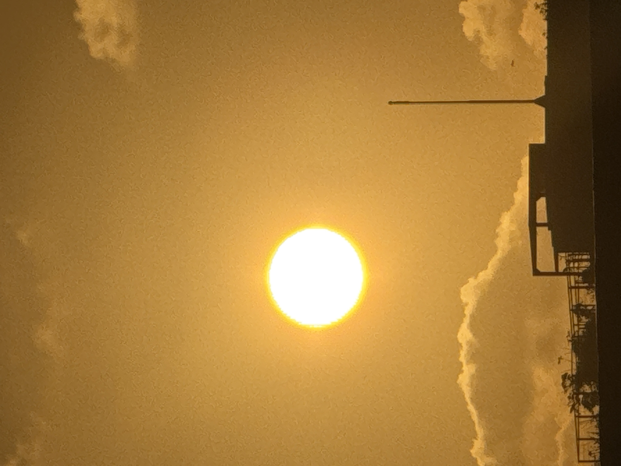 The image depicts a scene in Kuta Selatan, Indonesia, during what appears to be sunset or sunrise. A bright, luminous sun is positioned in the upper center of the frame, casting a warm, golden glow across the sky. The sky is filled with diffused light, with wisps of clouds scattered throughout, particularly along the horizon.
In the foreground, the silhouette of buildings and structures is visible. The primary structure appears to be a flat-roofed building with some architectural elements, possibly a balcony or overhang, protruding from its surface. To the right of this building, a tall, slender pole, likely an antenna or flagpole, stands prominently against the sky. Trees and other vegetation are silhouetted along the bottom edge of the image, suggesting a landscape with some greenery. The overall atmosphere is serene and peaceful, characterized by the warm light and the silhouetted forms.