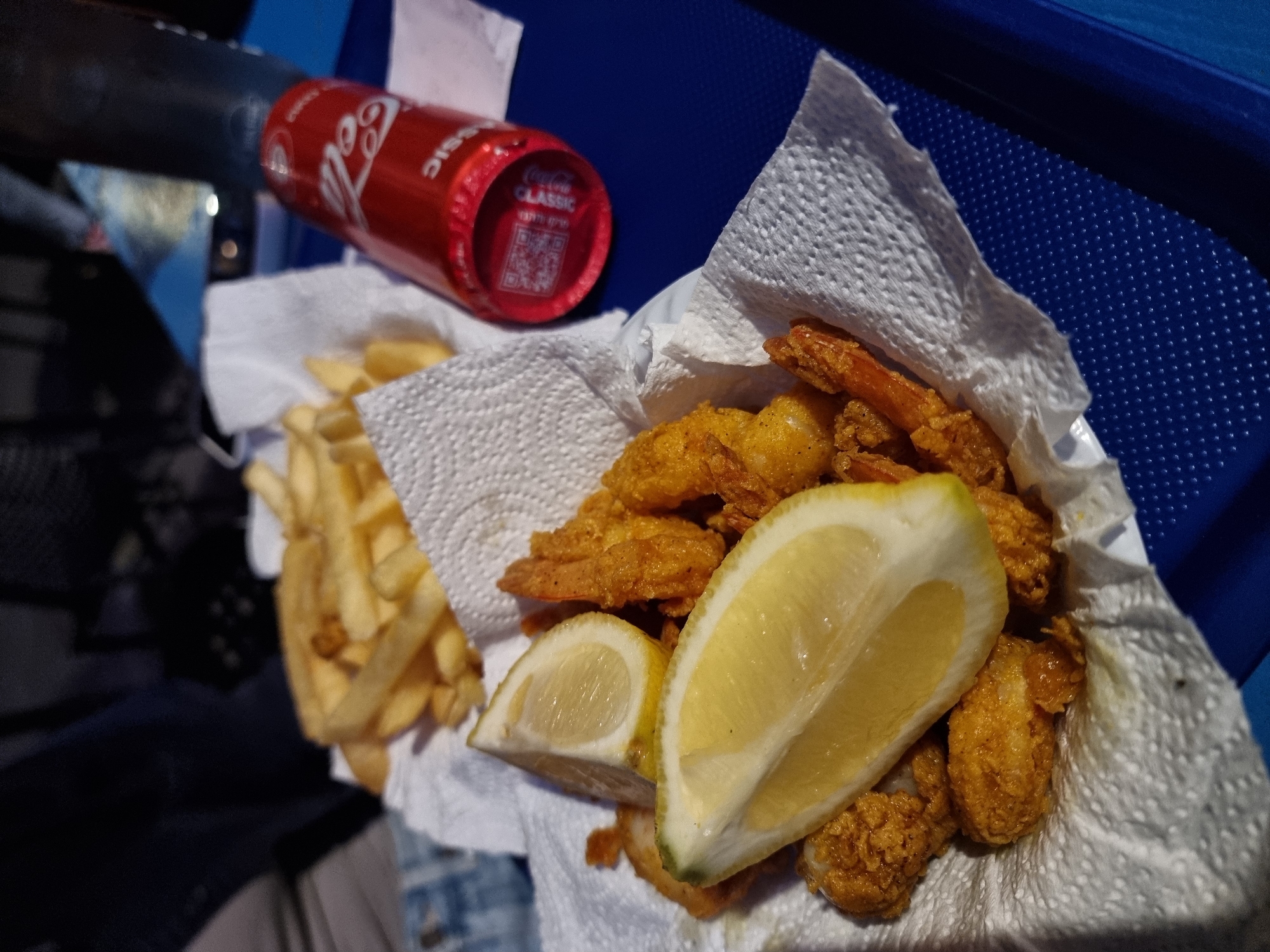 The image shows a meal of fried shrimp, french fries, and lemon wedges served on a white paper napkin on a blue tray. A can of Coca-Cola Classic is also present on the tray. The location and people are not discernible from the image. It's possible the image was taken in Tel-Aviv, Israel.