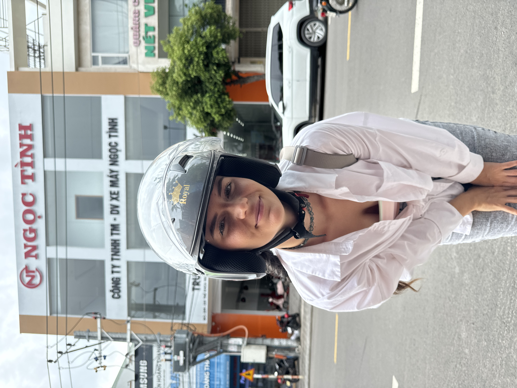A woman wearing a helmet sits in the street in Phu Thuy Ward, Vietnam. She is facing the camera with buildings and a parked white car behind her. A sign reads "NGỌC TÌNH," "CÔNG TY TNHH TM - DV XE MÁY NGỌC TÌNH" and "NÉT VI". Another sign reads "SAMSUNG" and "ĐÀNG TH." A utility pole with electrical wires is visible.
