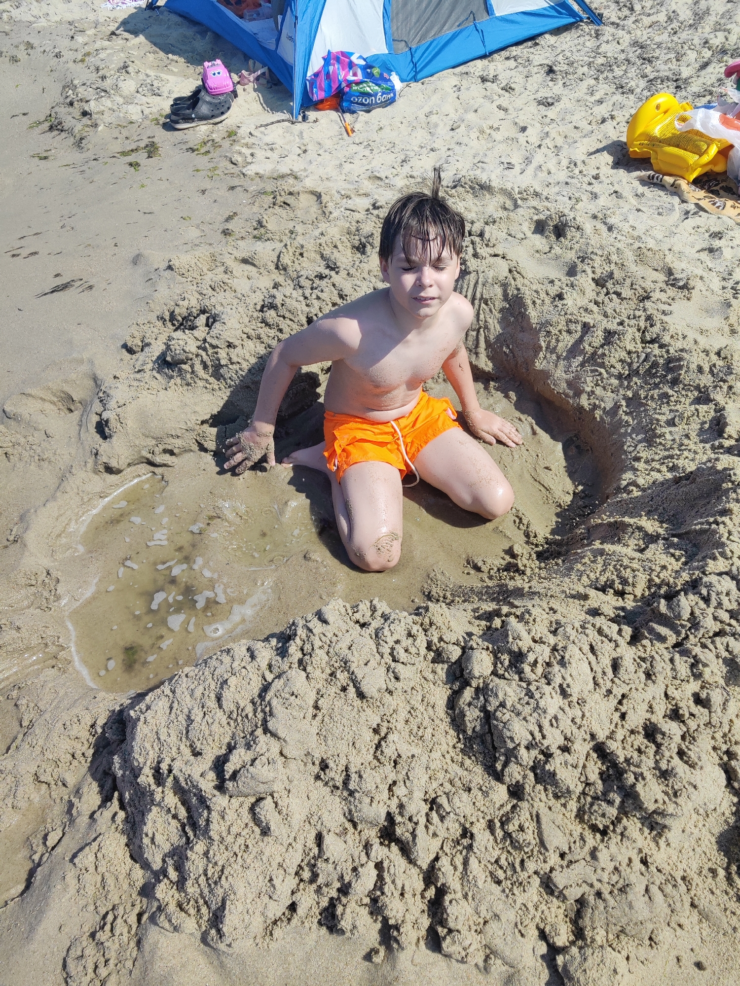 A young boy kneels in a hole dug in the sand on a beach. He is wearing orange swim trunks. The hole he is in has a small pool of water at the bottom. In the background, there is a blue and white tent, and beach toys. The location is Pionersky, Russia.