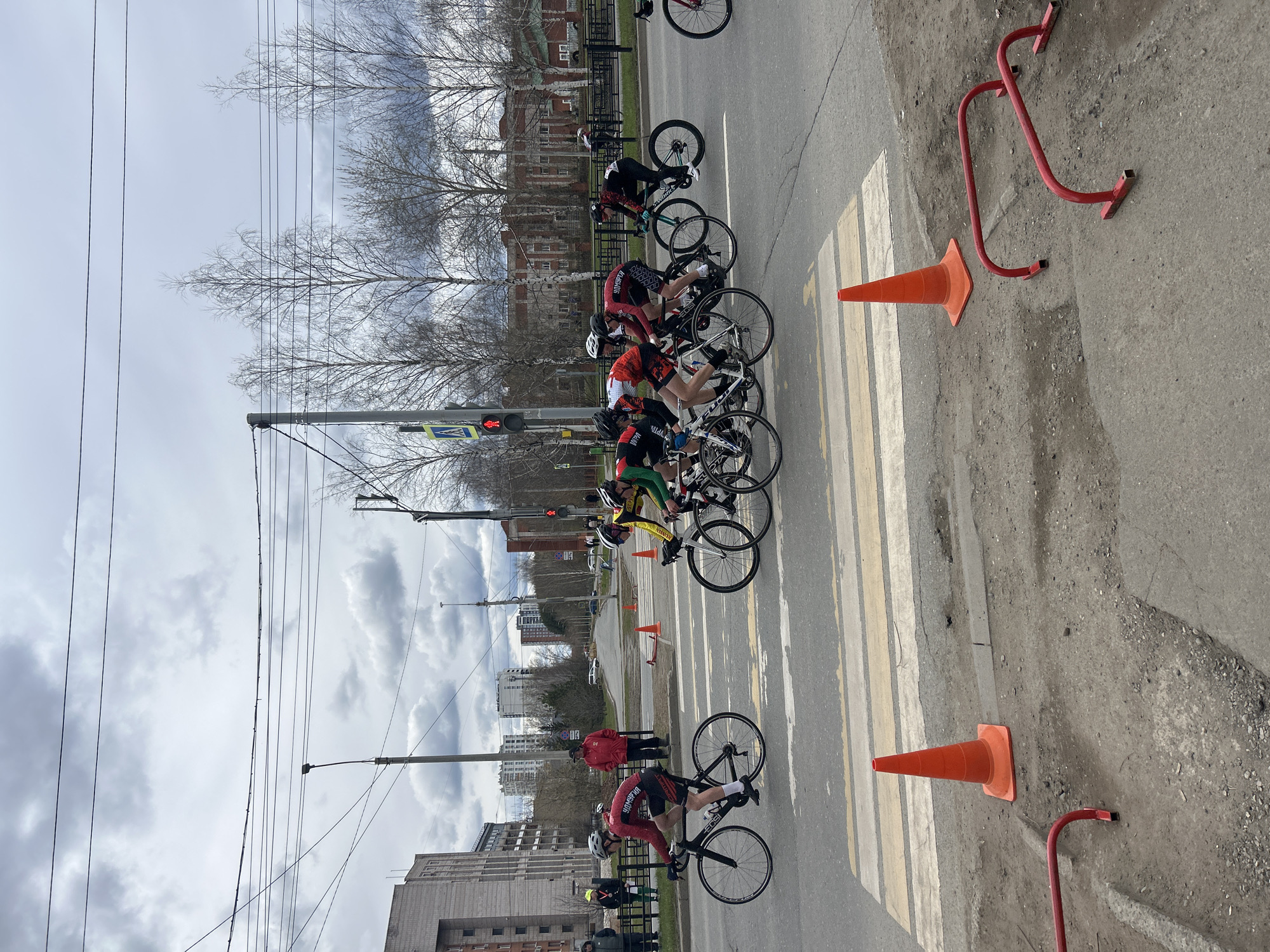 The image depicts a bicycle race in Izhevsk, Russia. Cyclists are positioned at a pedestrian crossing marked with white stripes. Orange cones delineate the course. A traffic light with a red signal is visible above the crossing. Buildings line the street, with bare trees in the background. Overhead power lines crisscross the sky. A man in a red jacket stands near the racers. A red metal barrier lies on the ground near a cone.