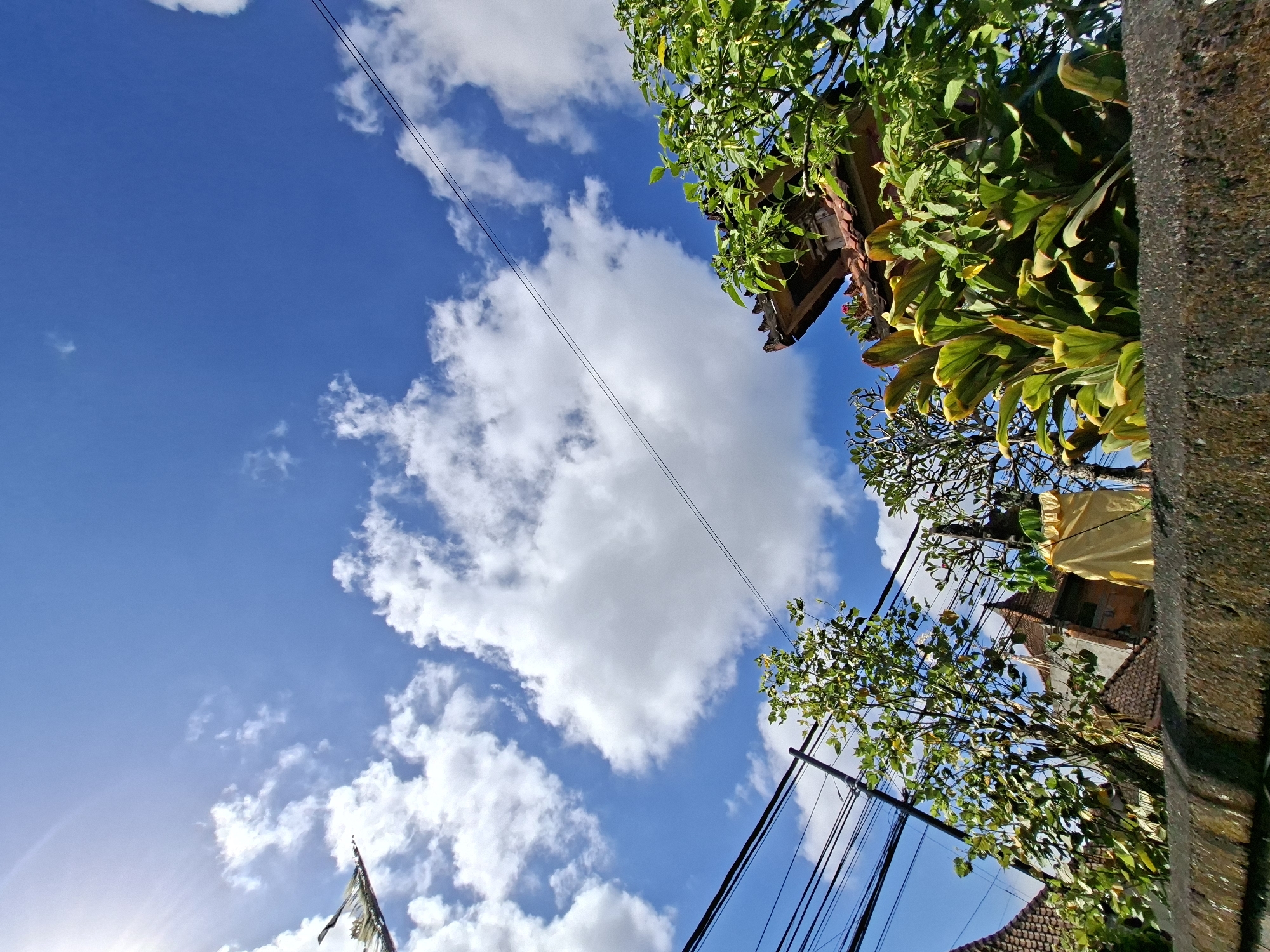 In Blahbatuh, Indonesia, a low-angle, wide shot captures a vibrant sky filled with fluffy white clouds against a deep blue backdrop. Sunlight streams in from the upper left, casting a bright glow. Several dark electrical wires traverse the sky, intersecting with the branches of lush green trees. In the foreground, a stone wall runs horizontally across the bottom of the frame. Beyond the wall, glimpses of traditional Indonesian architecture are visible, with terracotta tiled roofs and intricate building details. Large, leafy plants and trees with dense foliage are present, some adorned with bright yellow fabric. The scene conveys a sense of a bright, sunny day in a tropical environment with a blend of natural beauty and urban infrastructure.