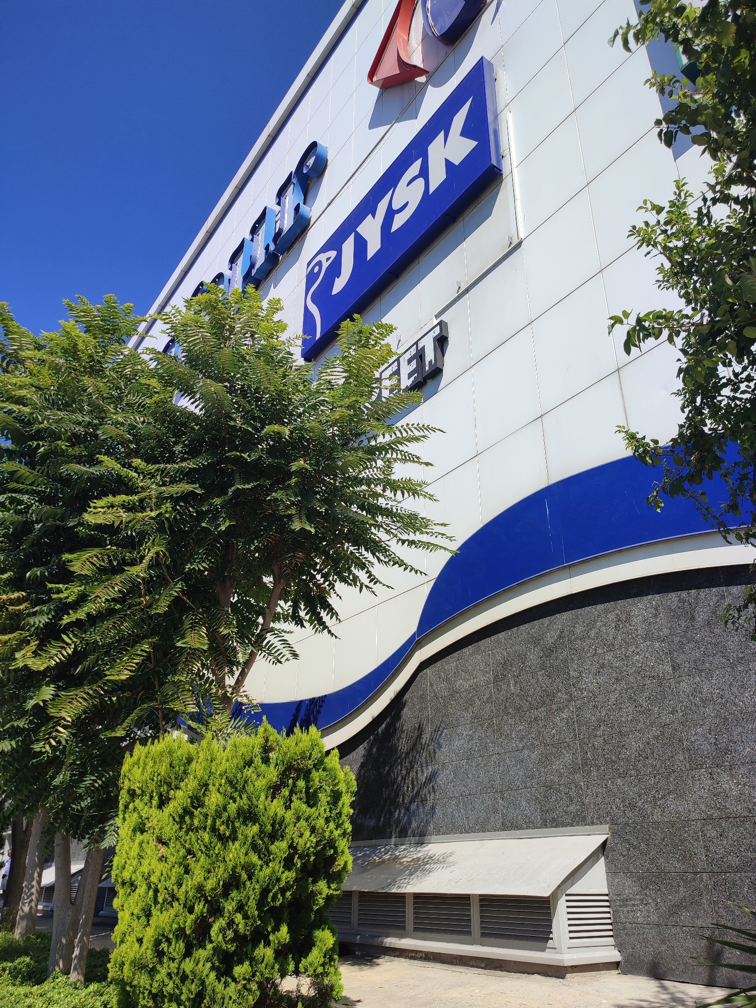 The image shows the facade of a modern building, likely a commercial establishment, in Kadıköy, Turkey. The building's exterior is a combination of white panels with blue accents and dark granite-like material on the lower section. Prominently displayed on the upper facade are large blue signs with white lettering. One sign reads "JYSK", a Scandinavian retailer. Another partially visible blue sign appears to be in a similar style. Above these, other blue letters are visible, suggesting additional branding or signage.
To the left of the building, lush green trees with feathery foliage are positioned in front of the facade. A more compact, dense green shrub is also visible in the foreground, to the left. On the right side of the building, more green foliage from trees partially obscures the view of the upper sections. The building's design includes a sweeping blue curved band that runs across the white facade, adding a dynamic visual element.
The lower part of the building features a dark, textured material, possibly granite tiles, arranged in a grid pattern. At the very bottom, a ventilation unit with louvers is set into the facade, indicating it's part of the building's infrastructure. The sky above is a clear, bright blue, suggesting a sunny day. The overall scene depicts an exterior view of a commercial building in an urban environment, likely in the district of Kadıköy, Turkey, given the context.