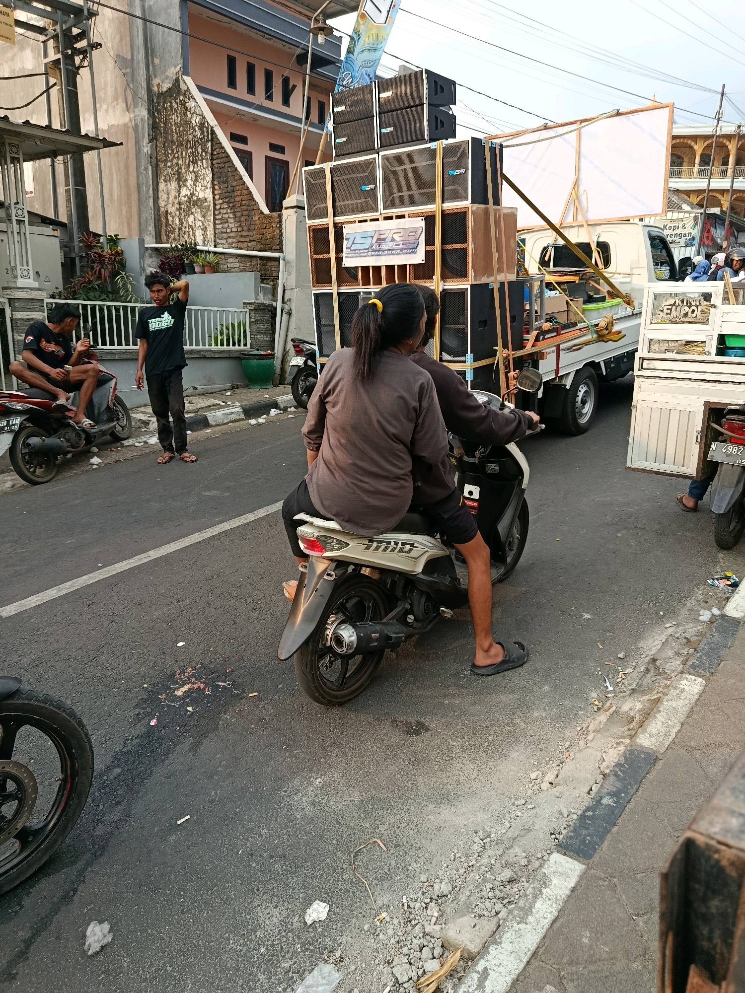 In Gubukklakah, Indonesia, an outdoor scene unfolds on a paved road in front of a multi-story building. The building, painted in shades of beige and pink, features dark-paned windows and a weathered, textured wall. In the foreground, two people ride a scooter. The rider, a woman with a yellow ponytail, wears a grey long-sleeved shirt and black shorts, while the passenger, a man, sports a black t-shirt and black shorts. They are positioned on the road, with a white scooter visible beneath them.
A large sound system, consisting of multiple stacked speakers, is mounted on a white truck, which is parked on the right side of the road. The truck also carries a large, white rectangular screen, supported by wooden framing. Another white truck with an open cargo area is parked behind the first one. Several people are visible near the trucks, including a man in a black t-shirt standing on the left side of the road, near two individuals seated on another scooter.
The road's surface is asphalt, with a distinct white line marking the lane. Debris, including paper scraps and what appears to be food waste, is scattered on the road. A storm drain with a broken edge is present along the curb. A black motorcycle is parked on the far left, its front wheel visible. The overall atmosphere suggests a street scene, possibly a temporary setup for an event or gathering.