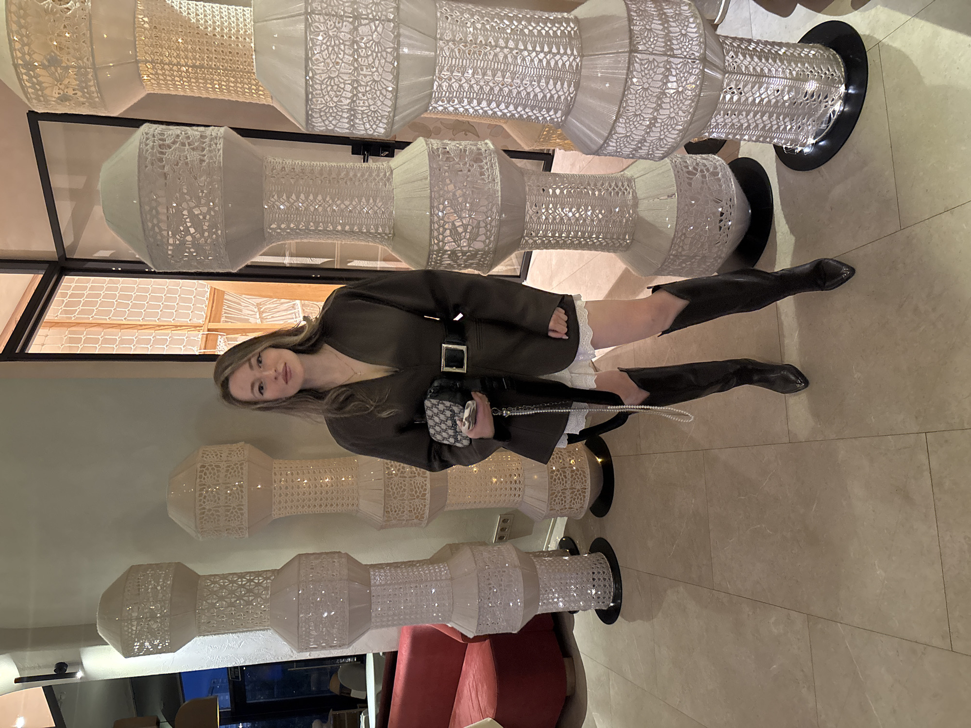 The image shows a woman posing indoors, likely in a commercial space. She is wearing a dark green jacket with a belt, a white skirt, and black over-the-knee boots. She is carrying a small black and white patterned bag with a pearl strap. The background features a row of decorative lamps with a white lace-like pattern, mounted on black bases. A partial view of a red sofa is visible in the bottom left corner. The floor is light-colored tile. The location is unspecified, and there is no indication that the photo was taken in Krasnoyarsk, Russia.