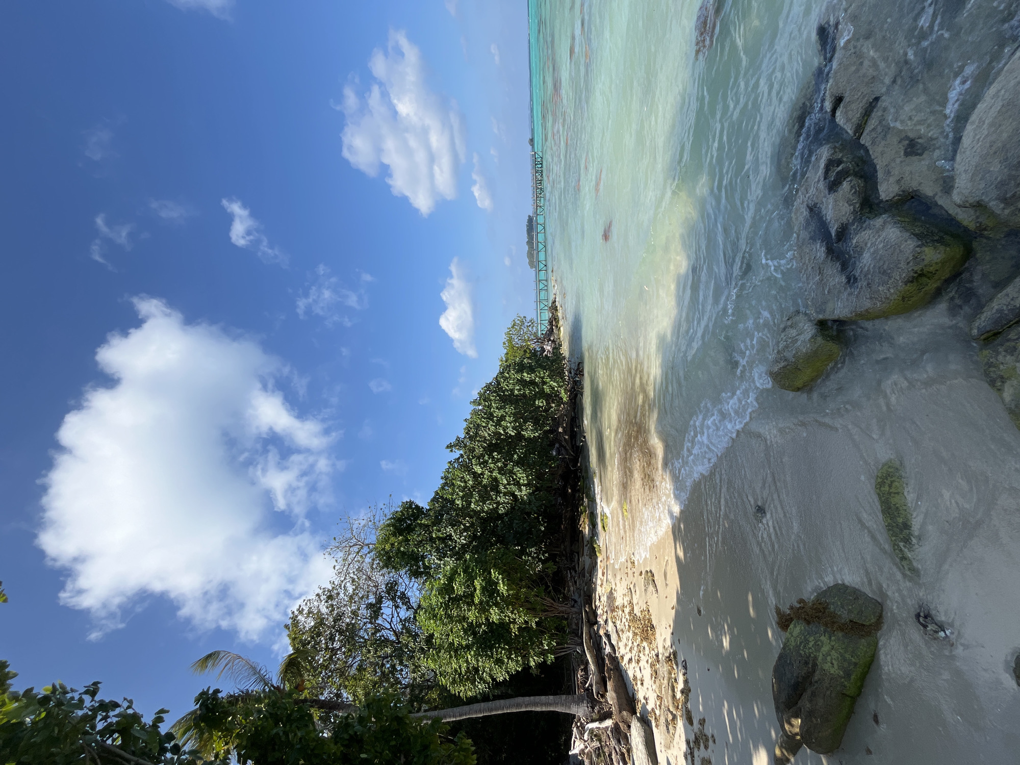 This is a daytime, eye-level, medium shot of a tropical beach in Legonipah, Indonesia. The scene features a sandy shoreline with scattered rocks and driftwood, leading to clear turquoise water. Lush green trees line the back of the beach. In the distance, a wooden pier extends into the ocean, connecting to a small island. The sky is bright blue with scattered white clouds. There are no people or animals visible in the image.