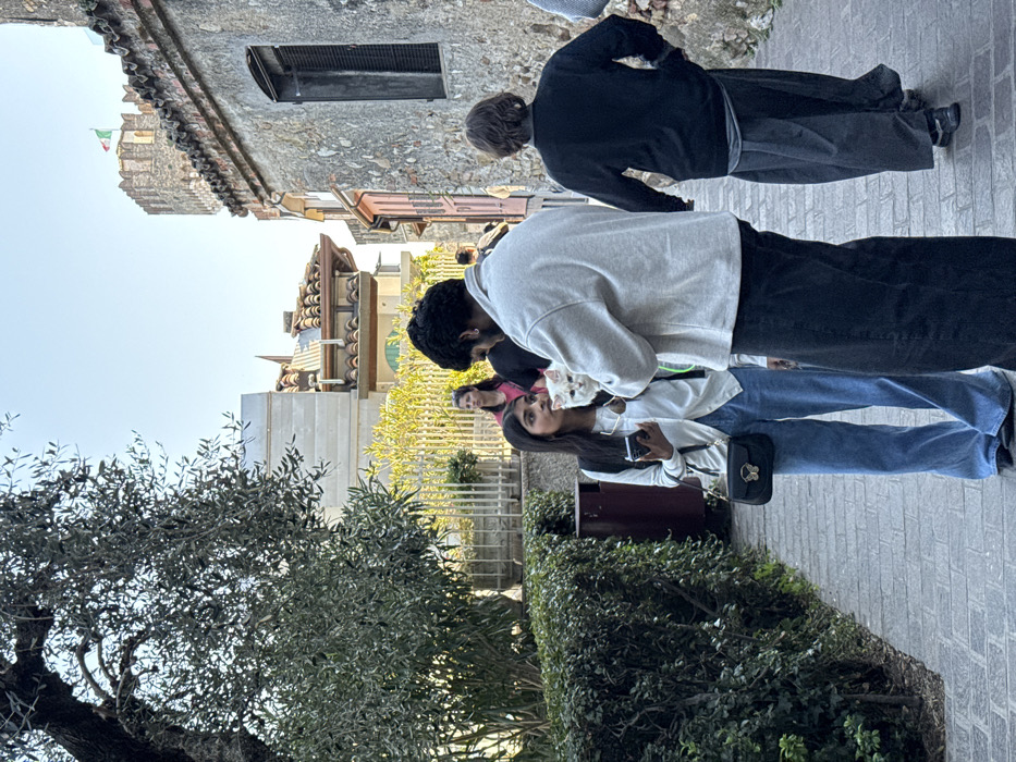 The image displays an outdoor scene on a paved pathway in Sirmione, Italy. In the foreground, a young adult female with dark hair stands on the left, wearing a white cardigan, blue wide-leg jeans, and a black shoulder bag with a gold butterfly clasp; she holds a white smartphone. Opposite her, a young adult male with dark curly hair, seen mostly from the back, wears a light grey long-sleeved top and dark trousers; he is holding a white long-haired cat. The cat is facing towards the female. To the right, an individual with dark hair, dressed in a dark top and wide dark trousers, walks away from the camera. In the midground, a female in a pink top is partially visible behind a white metal railing and green foliage on the left. The pathway is flanked by dense green hedges and an olive tree on the left, and old stone buildings with terracotta tiled roofs and dark window shutters on the right. A section of a light pink building facade is visible further down the street. In the upper right background, a crenellated castle tower, identifiable as part of Scaliger Castle, flies an Italian flag against a clear blue sky.