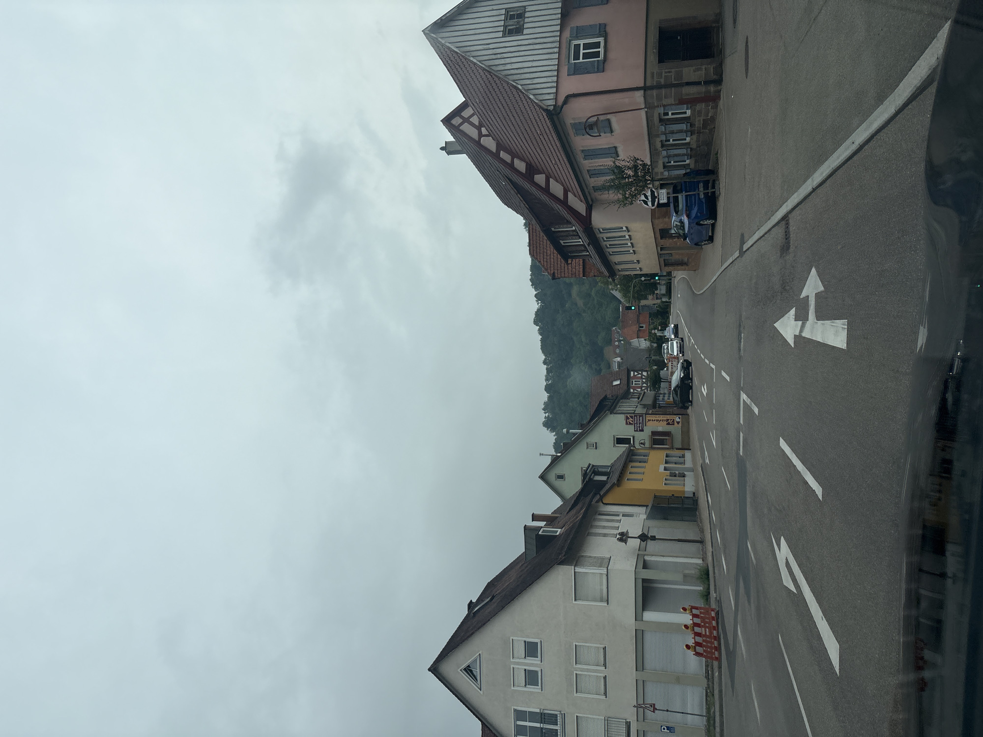 This image captures a street scene in Untermünkheim, Germany, under an overcast sky. The perspective is from inside a vehicle, looking forward and slightly to the right. The road features lane markings, including a white arrow indicating a turn to the left or go straight, and another arrow pointing to the right.
On the left side of the street, there is a light-colored building with multiple windows and closed shutters. Further down the street on the left, there's a yellow building and other structures with reddish-brown roofs, consistent with traditional German architecture. Orange construction barriers are visible on the sidewalk in front of the light-colored building.
On the right side of the street, a building with a pink facade and a red-tiled roof is prominent. This building has several windows, some with blue shutters. A street lamp is positioned in front of it. A blue car is parked on the right side of the road, behind a small tree.
In the background, a wooded hill rises, suggesting the town is situated in a valley or near a natural landscape. Several cars are visible on the road in the distance. The overall impression is a quiet, residential or mixed-use street in a German town.