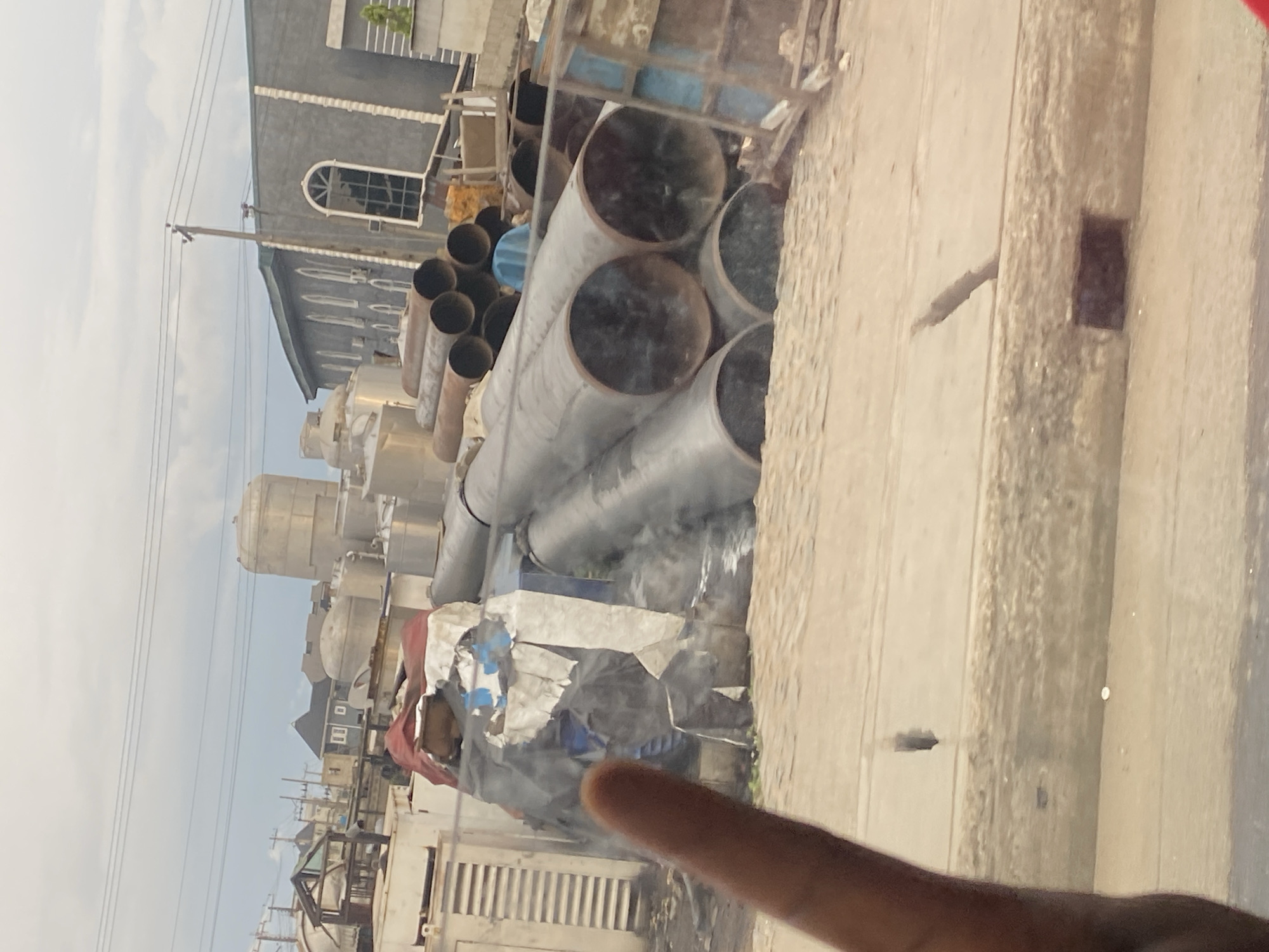 The image depicts an outdoor scene in Ikeja, Nigeria, featuring a collection of large metal pipes stacked on the ground. Some of the pipes are covered with tarpaulin and sheeting. In the background, there are buildings, including what appears to be a church with arched windows and other industrial structures with tanks. Overhead, power lines are visible against a cloudy sky. The foreground shows a dirt or gravel surface with a partial view of a person's finger on the left side of the frame, suggesting the photo was taken from inside a vehicle or through a window. The overall environment suggests an industrial or construction area.