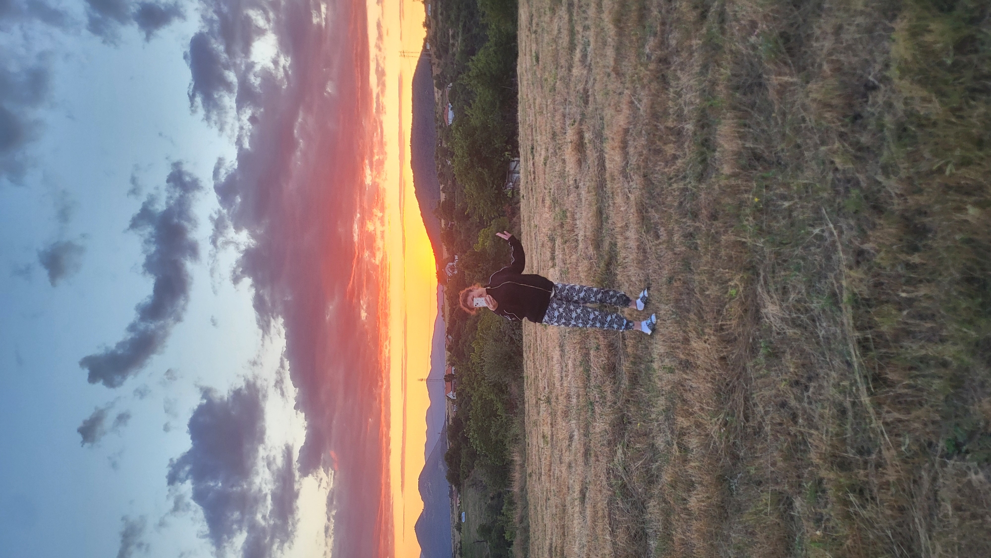 A full-body, eye-level, outdoor, vertical shot captures a young person standing in a dry, grassy field during sunset in the city of Rechitsa, Bulgaria. The person, with curly auburn hair, wears a black zip-up hoodie, camouflage-patterned pants, white socks, and black slides. They are holding a white smartphone in their right hand, presumably taking a selfie, and their left arm is extended outwards. The background features rolling hills covered in green trees, dotted with houses and a cell tower, all silhouetted against a vibrant sunset sky. The sky transitions from a pale blue at the top to fiery oranges and reds near the horizon, with scattered dark clouds. The sun is a bright orange orb just above the horizon, casting a warm glow over the landscape. The foreground is dominated by the dry, golden-brown grass, indicating a summer or early autumn setting.