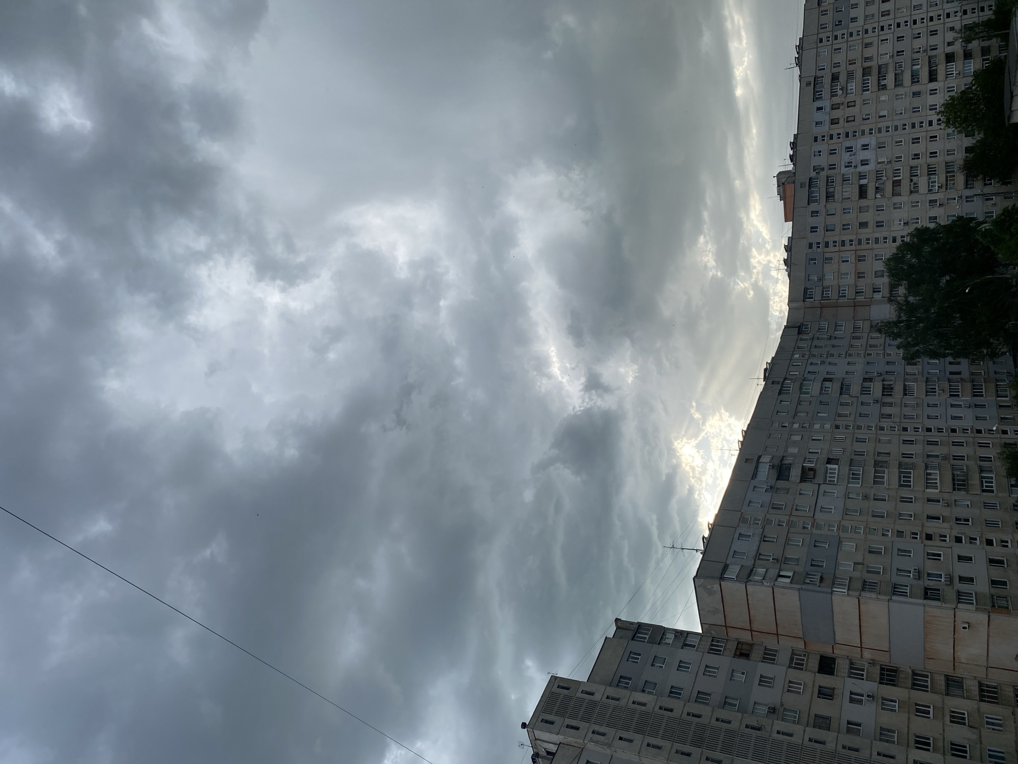 The image shows a low-angle view of two high-rise apartment buildings in Kharkiv, Ukraine, under a sky filled with heavy, dark clouds. The buildings appear to be of Soviet-era construction, characterized by repetitive, rectangular window patterns and a generally utilitarian architectural style. The sky is predominantly gray, with hints of sunlight breaking through the clouds in the upper portion of the frame. A power line stretches across the lower left portion of the image, contrasting with the sky. The overall scene conveys a sense of urban density and the potential for inclement weather.