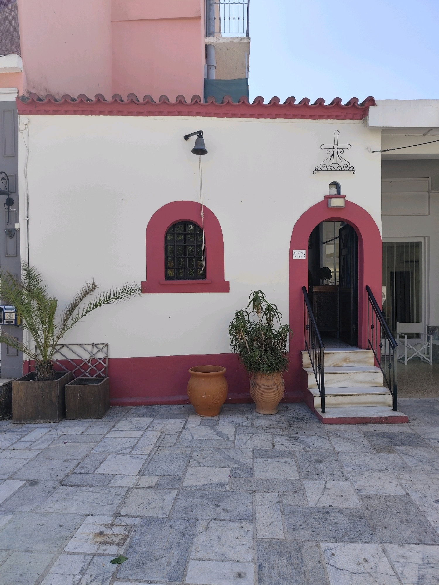 The image depicts the exterior of a small building, likely a chapel or a similar structure, with a distinct architectural style. The building features a white facade with a red-tiled roof and red trim around the arched entryway and a rectangular window. Above the window, a black bell is mounted on the wall. To the right of the entrance, an ornate metal cross is affixed to the facade.
The entrance is an arched doorway with stairs leading up to it. The doorway is framed in red, and a sign is visible on the wall to the left of the entrance. The stairs are made of marble with black metal railings.
In front of the building, on the paved ground, there are several potted plants. To the left, a palm tree is planted in a wooden planter, and next to it are two more wooden planters. To the right of the stairs, a large potted plant with green foliage is situated. Two terra cotta pots are also visible on the ground. The ground itself is paved with large, irregularly shaped stone tiles.
In the background, parts of other buildings are visible, including a pink-colored structure. The sky is clear and blue. The scene appears to be set in a Mediterranean environment, possibly a village or town.
Based on the provided information, this location is in the Municipal Unit of Nafplio, Greece.
