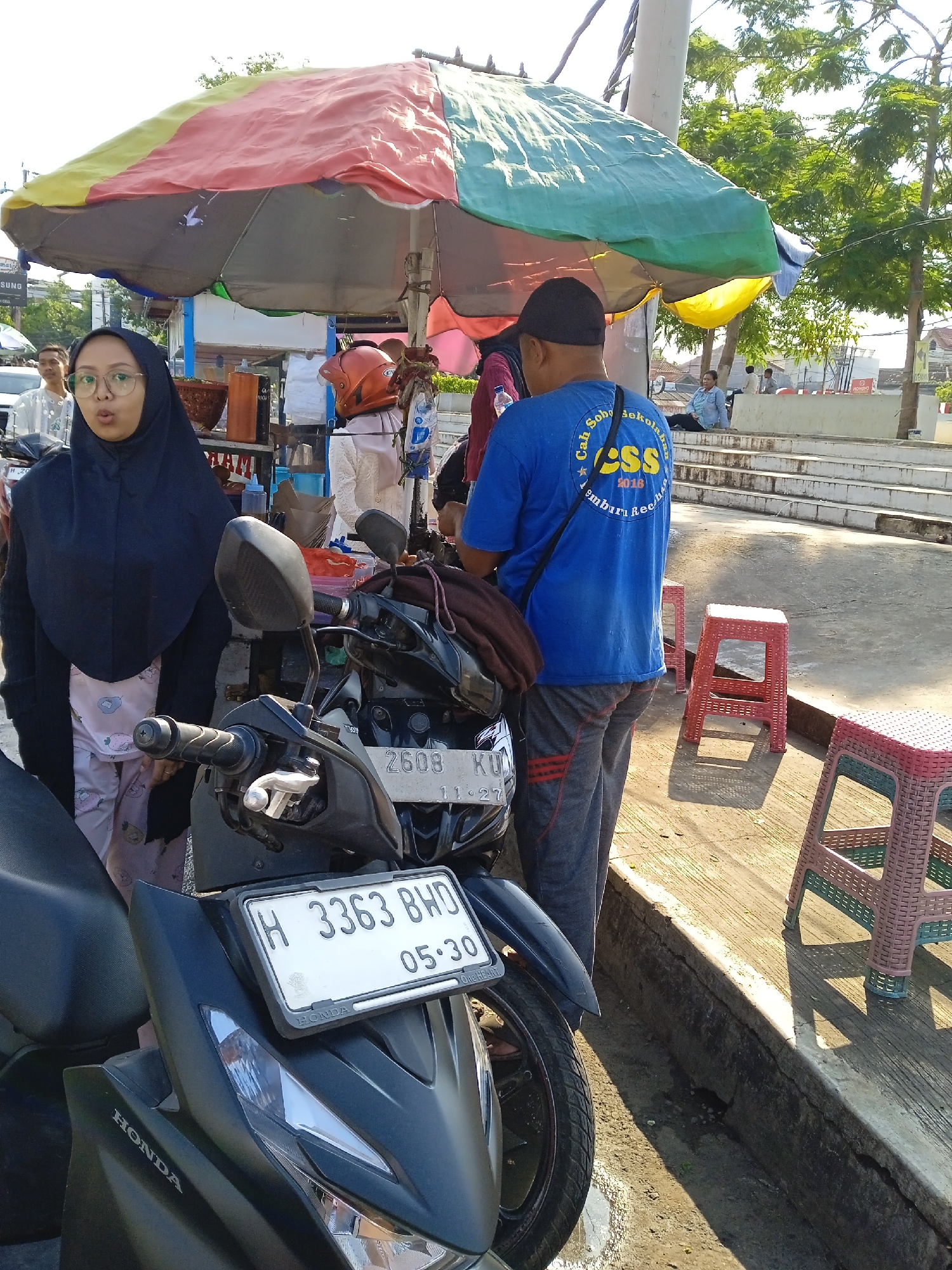 The scene depicts a street food vendor in Kaliwungu, Indonesia, based on license plate identification. A woman wearing a hijab and glasses stands beside a black Honda motorcycle, license plate "H 3363 BWD 05-30". Another motorcycle is parked nearby, license plate "2608 KU 11-27". A man wearing a blue t-shirt with "CSS 2016" printed on the back is standing at the vendor stall. The vendor stall is covered by a colorful umbrella. There are several red plastic stools near the stall. Other people are sitting on steps in the background.