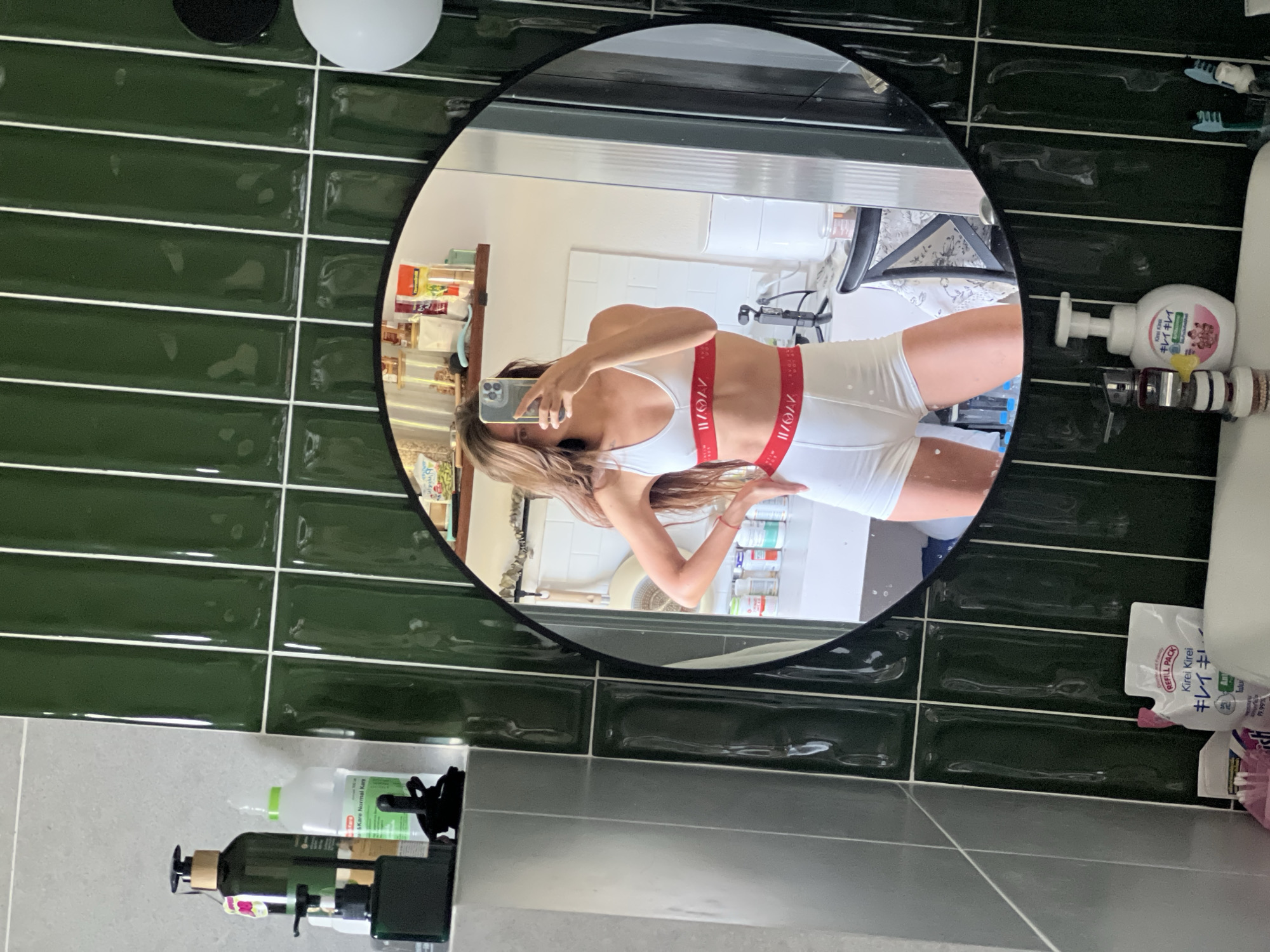 A mirror reflects a person taking a selfie in a bathroom. The person is wearing a white top and bottoms with red trim. The bathroom features green subway tiles, a white toilet, and various toiletries.