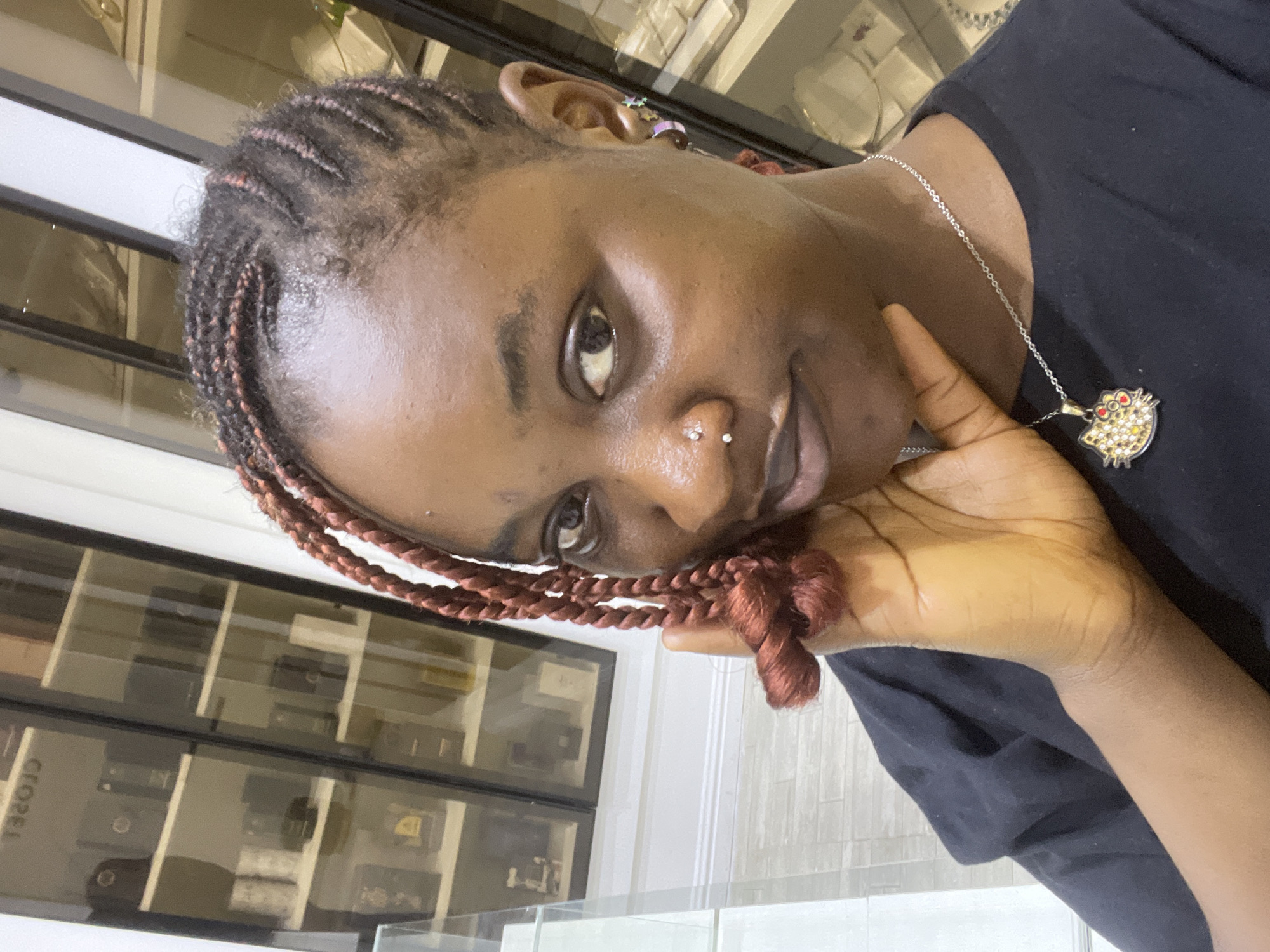 The media file is a portrait of a young Black woman indoors. She is wearing a black t-shirt and a silver necklace with a pendant shaped like a pufferfish. Her hair is styled in braids with reddish-brown extensions. She has a nose ring and multiple earrings. Her right hand is resting on her cheek, and she is looking directly at the camera with a slight smile. The background appears to be a retail or display area with glass shelves holding various items, suggesting a shop or store. The lighting is bright, indicating it is daytime. The image is oriented vertically.